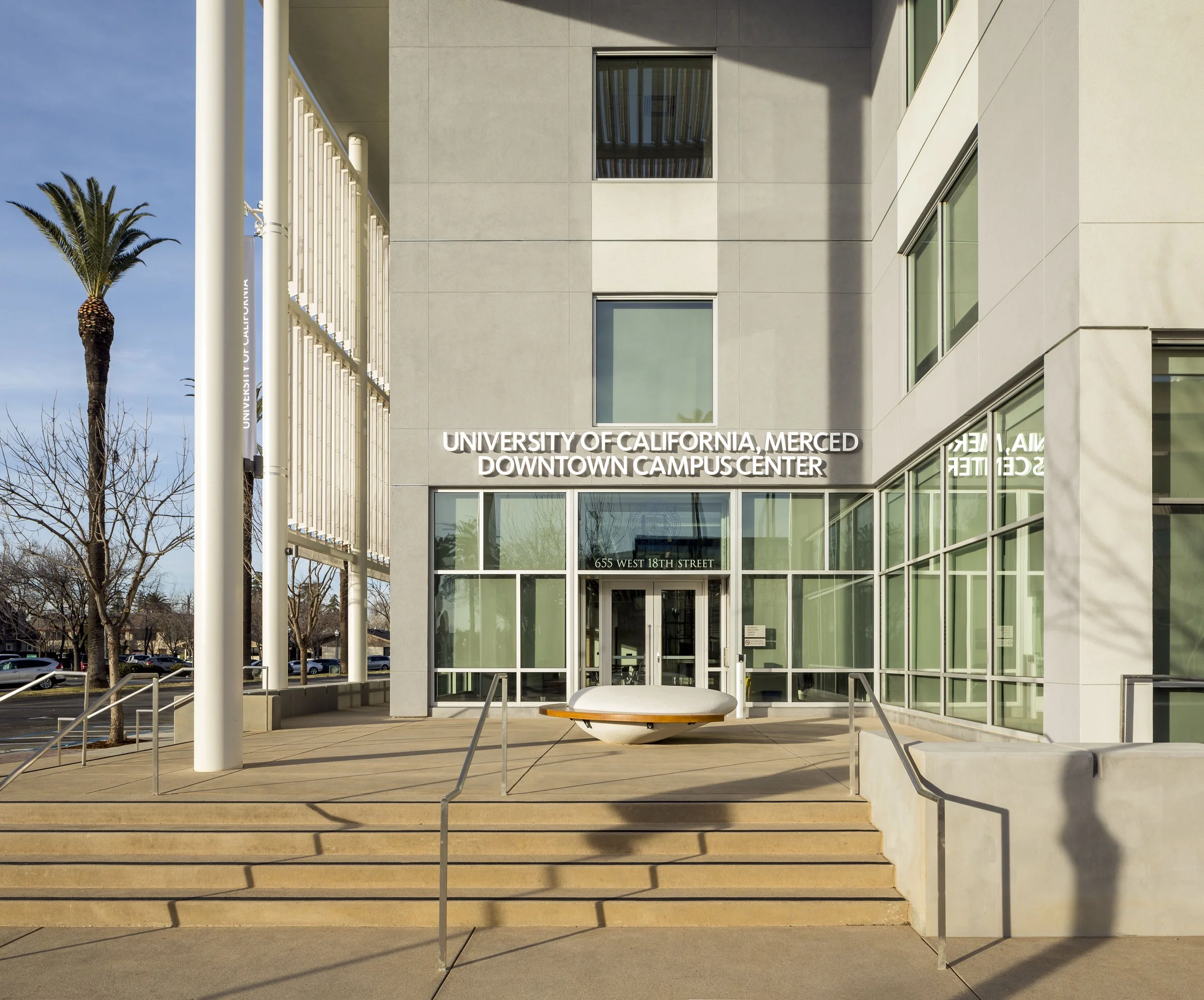 Facade of the University of California, Merced Downtown Campus Center with steps leading to glass entrance doors, palm trees, and a statue in the foreground.