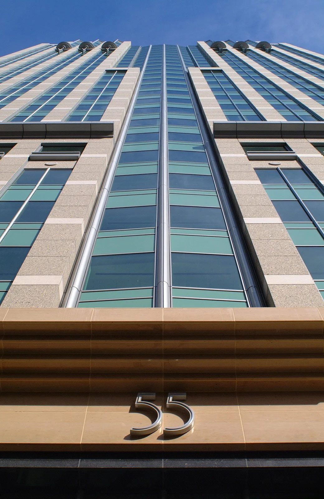 Low-angle view of a modern high-rise building with the number 55 displayed near the entrance and a clear blue sky overhead.