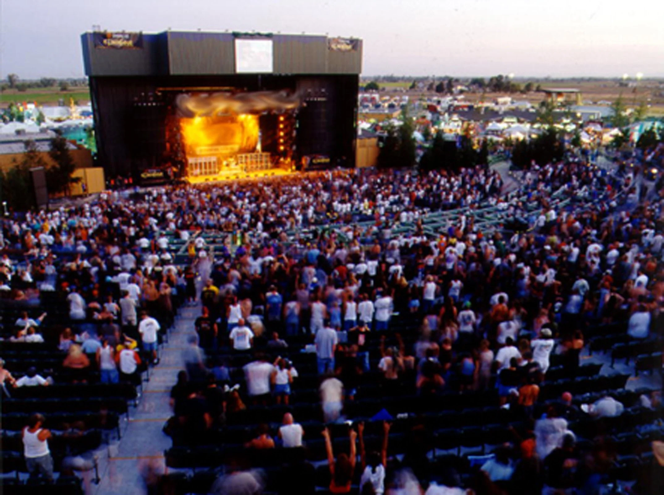 Large outdoor concert with a stage, flames, and many audience members seated in front.