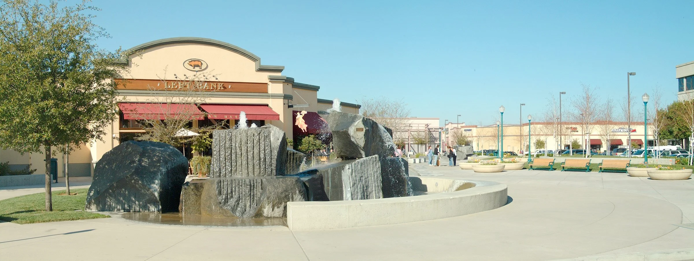 Outdoor shopping mall plaza with a fountain, trees, benches, and retail stores in the background on a clear day.