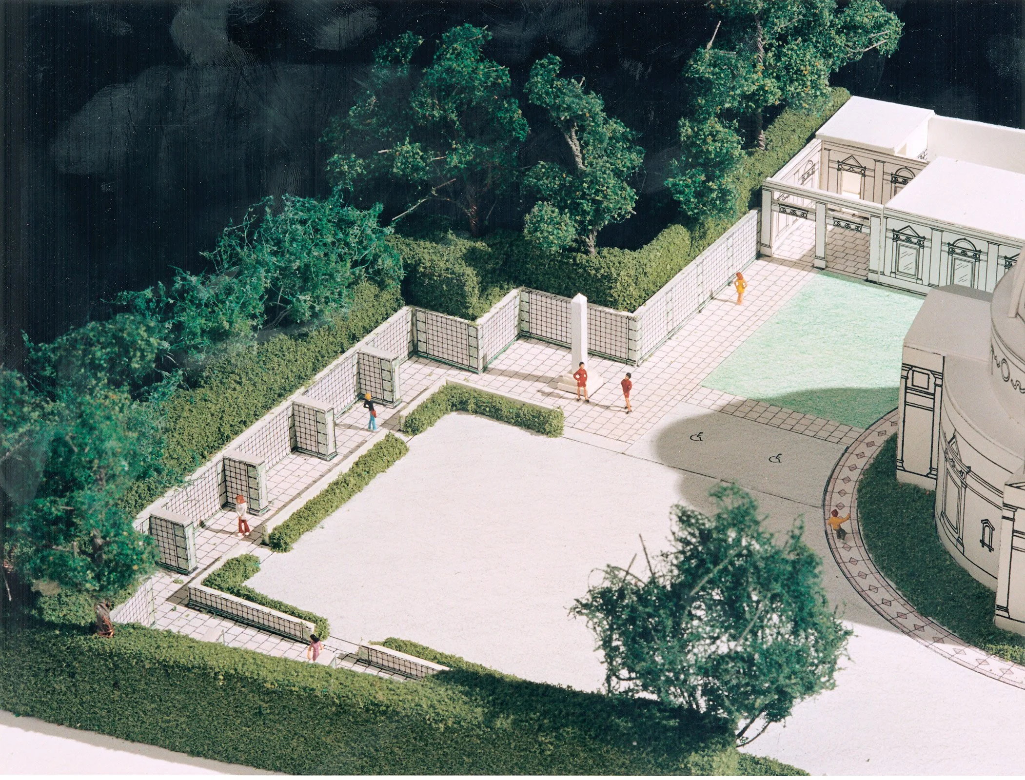 Miniature architectural model of a building with a courtyard, surrounded by trees and hedges, featuring small figurines of people walking and a tiled pathway.