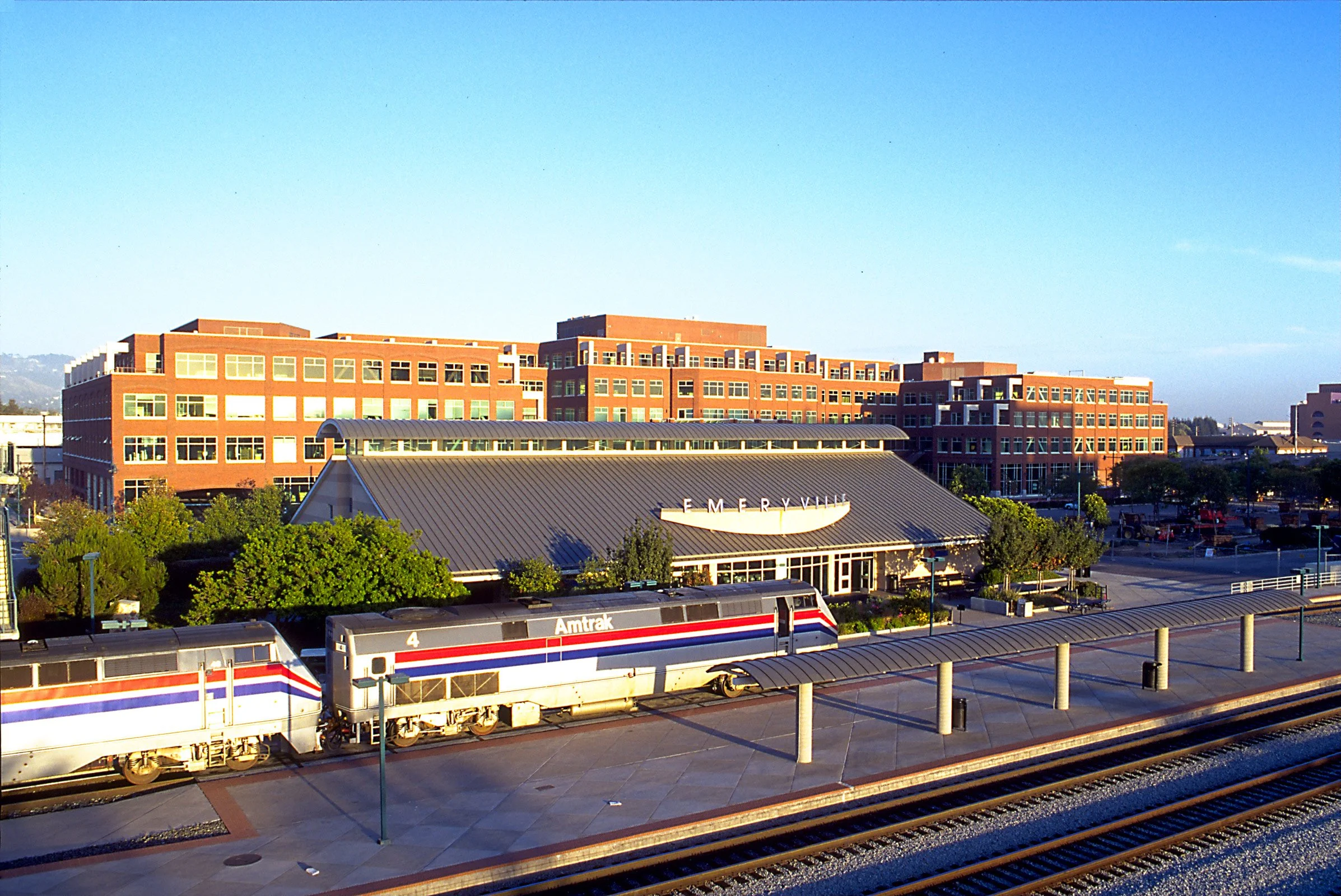 Amtrak train stopped at a station with the Emeryville sign, and a large brick building in the background under a clear blue sky.