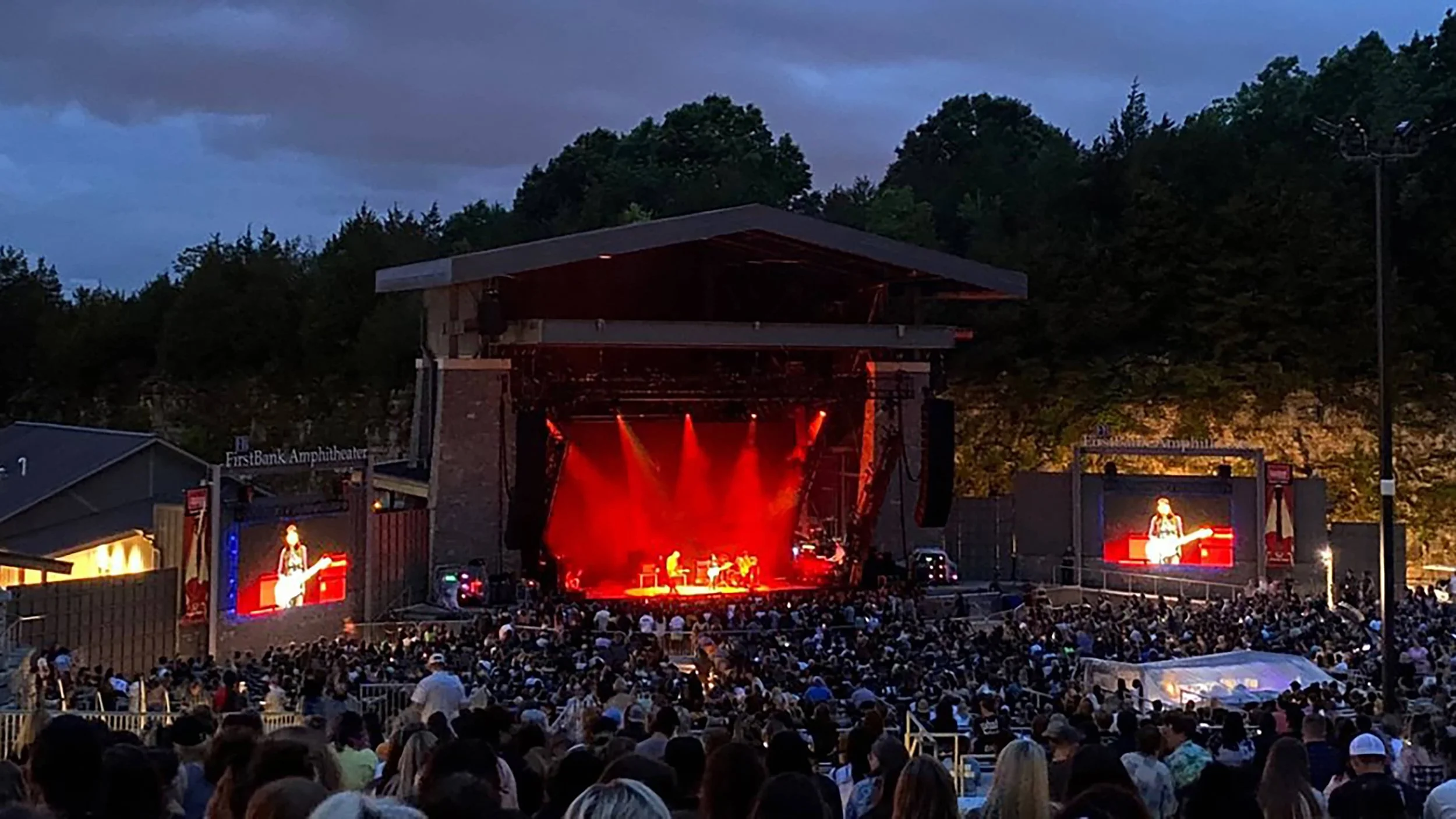 Concert at an outdoor amphitheater with a large audience, performers on stage illuminated by red lights, large screens displaying the performers, surrounded by trees and evening sky.