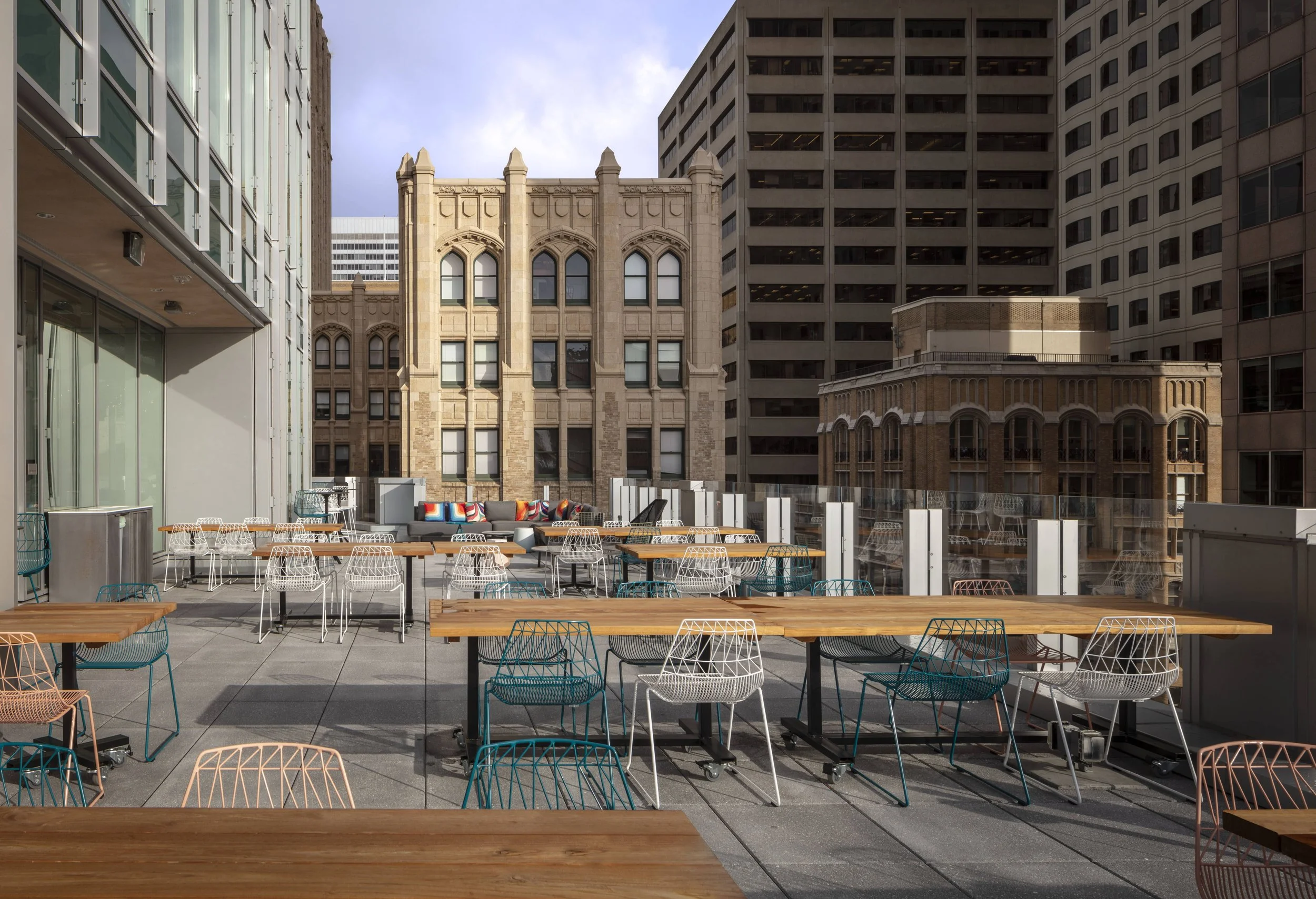 Empty outdoor rooftop patio with wooden tables and colorful wire chairs, high-rise buildings in the background, partly cloudy sky.