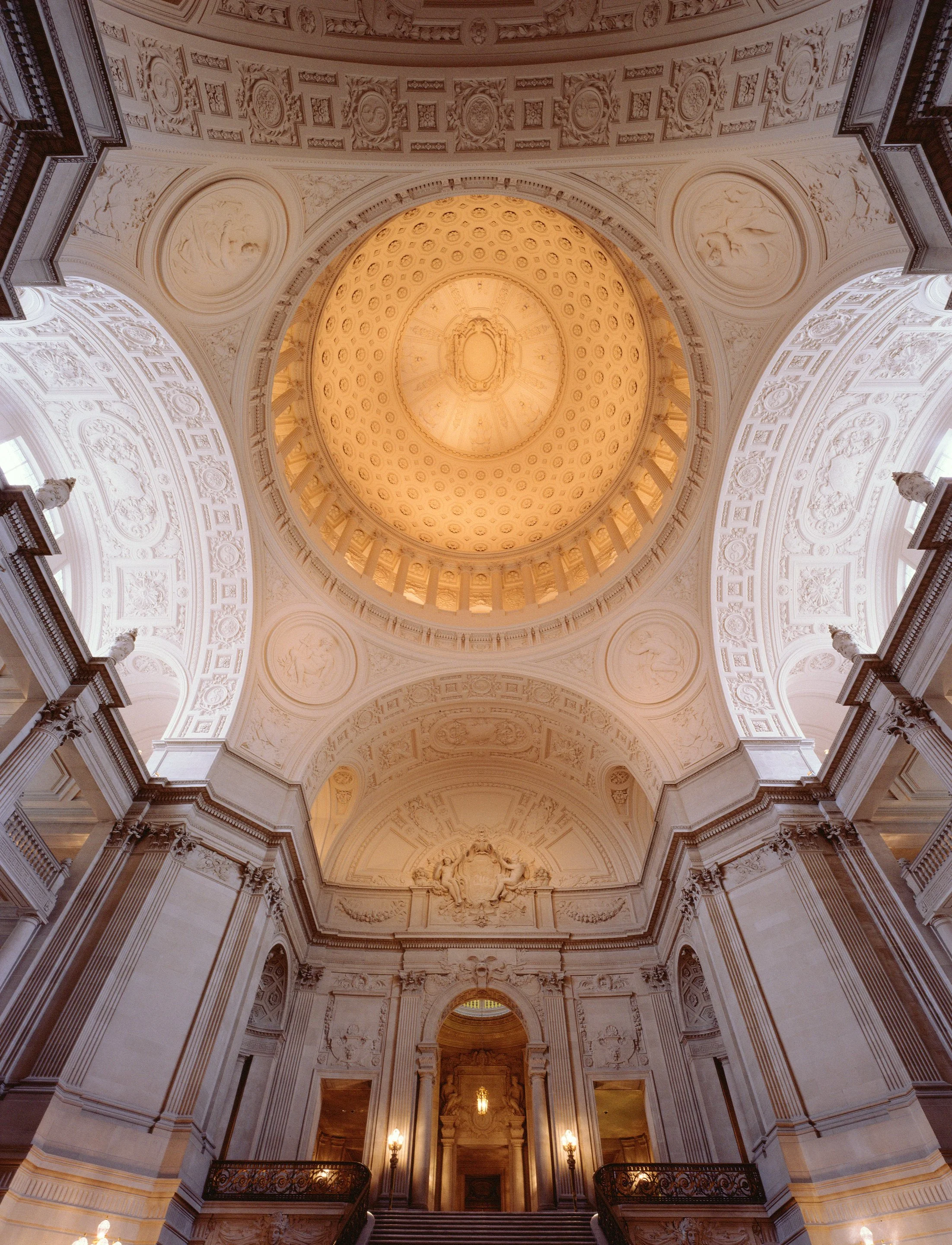 The interior view of a grand, ornately decorated building with a large, illuminated dome ceiling featuring intricate patterns and sculptures, and tall columns and staircase leading to a doorway.
