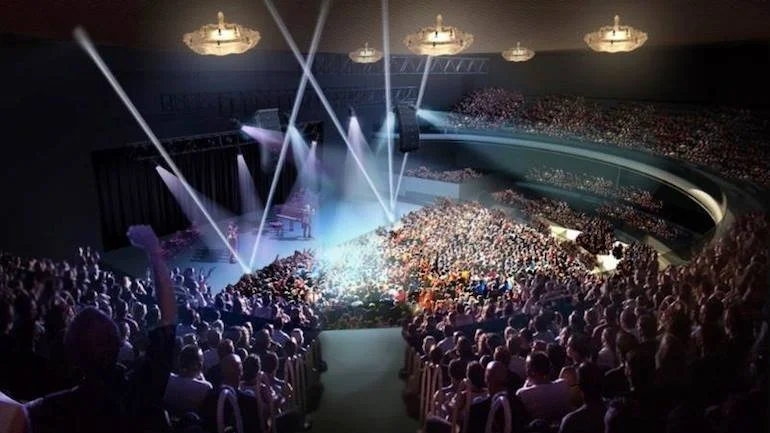 Large indoor concert venue filled with a crowd, stage with musicians, bright lighting, and spotlights shining on the stage.