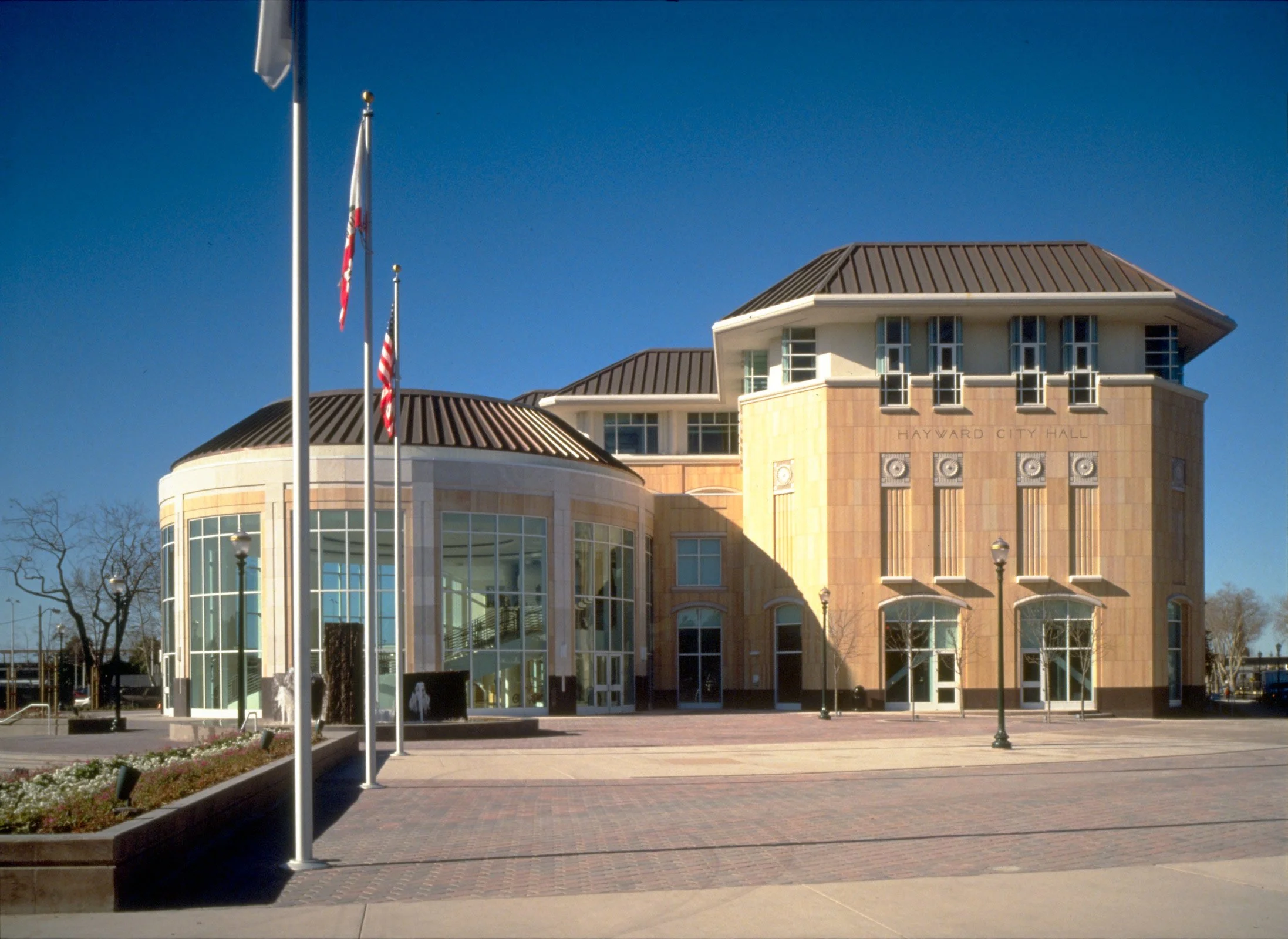 Exterior of Hayward City Hall building with flags and trees against a clear blue sky.