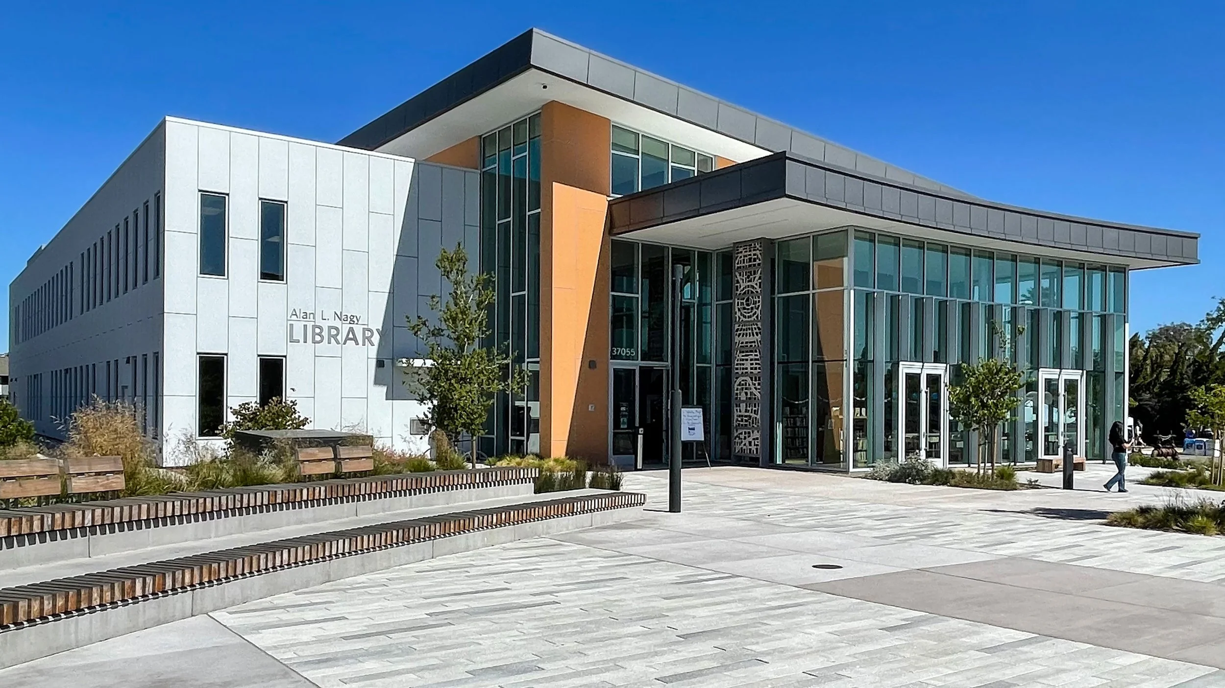 Modern library building with large glass windows and a landscaped front area with benches and small trees.