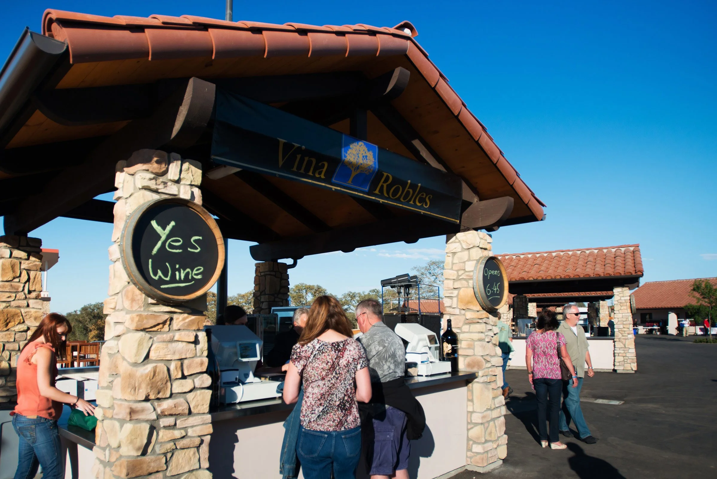 An outdoor wine tasting or purchase booth with stone pillars and a wooden roof underside. Signage indicates 'Yes Wine' and 'Opens 6:45.' People are gathered around the counter, and the sky is clear and blue.
