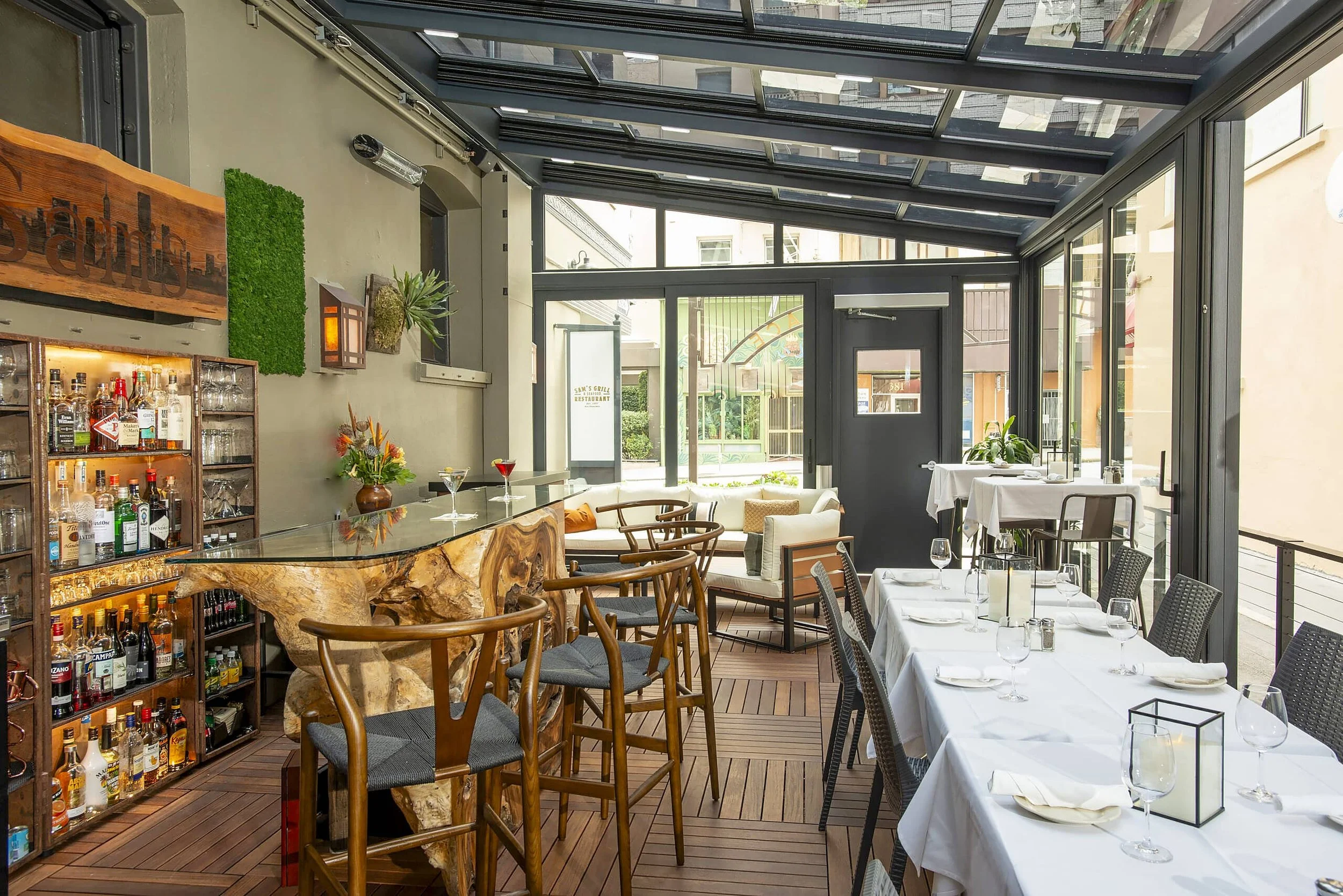 Interior of a restaurant with large windows, a bar with a wooden top, a shelf with liquor bottles, tables set with glasses and napkins, and a cozy seating area with a couch and armchairs.