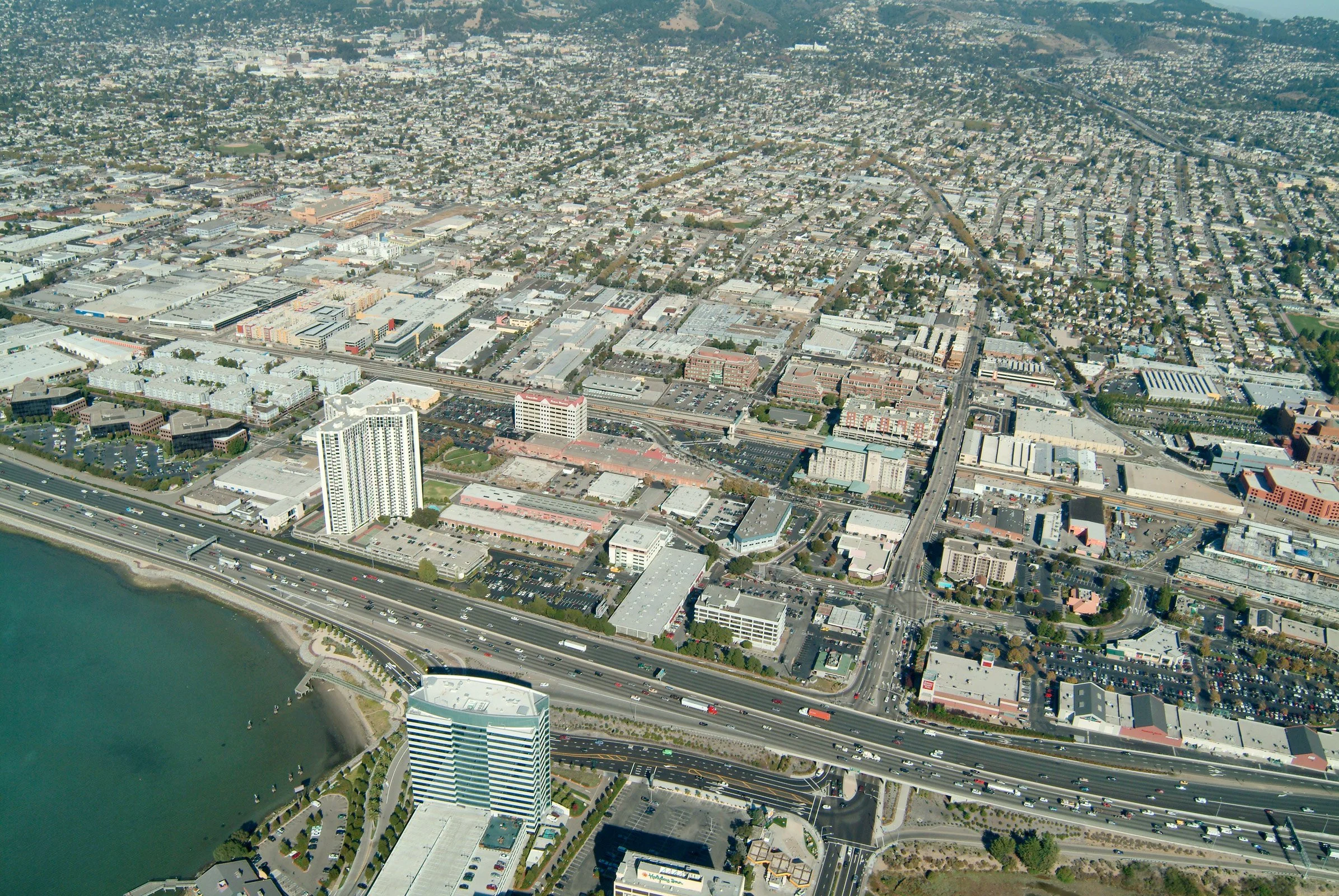 An aerial view of a cityscape showing a waterfront, high-rise buildings, a highway, and dense city blocks with residential and commercial buildings.