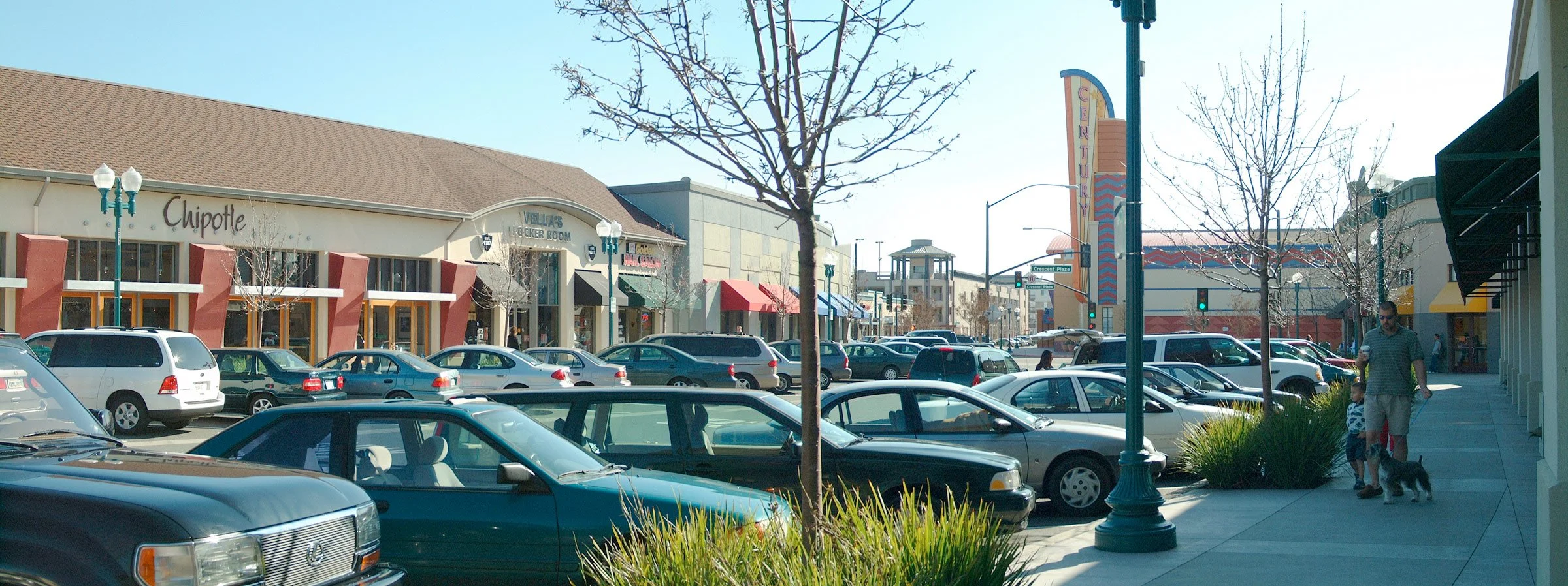 A busy outdoor shopping center on a sunny day with parked cars, leafless trees, storefronts including Chipotle, and people walking with dogs.