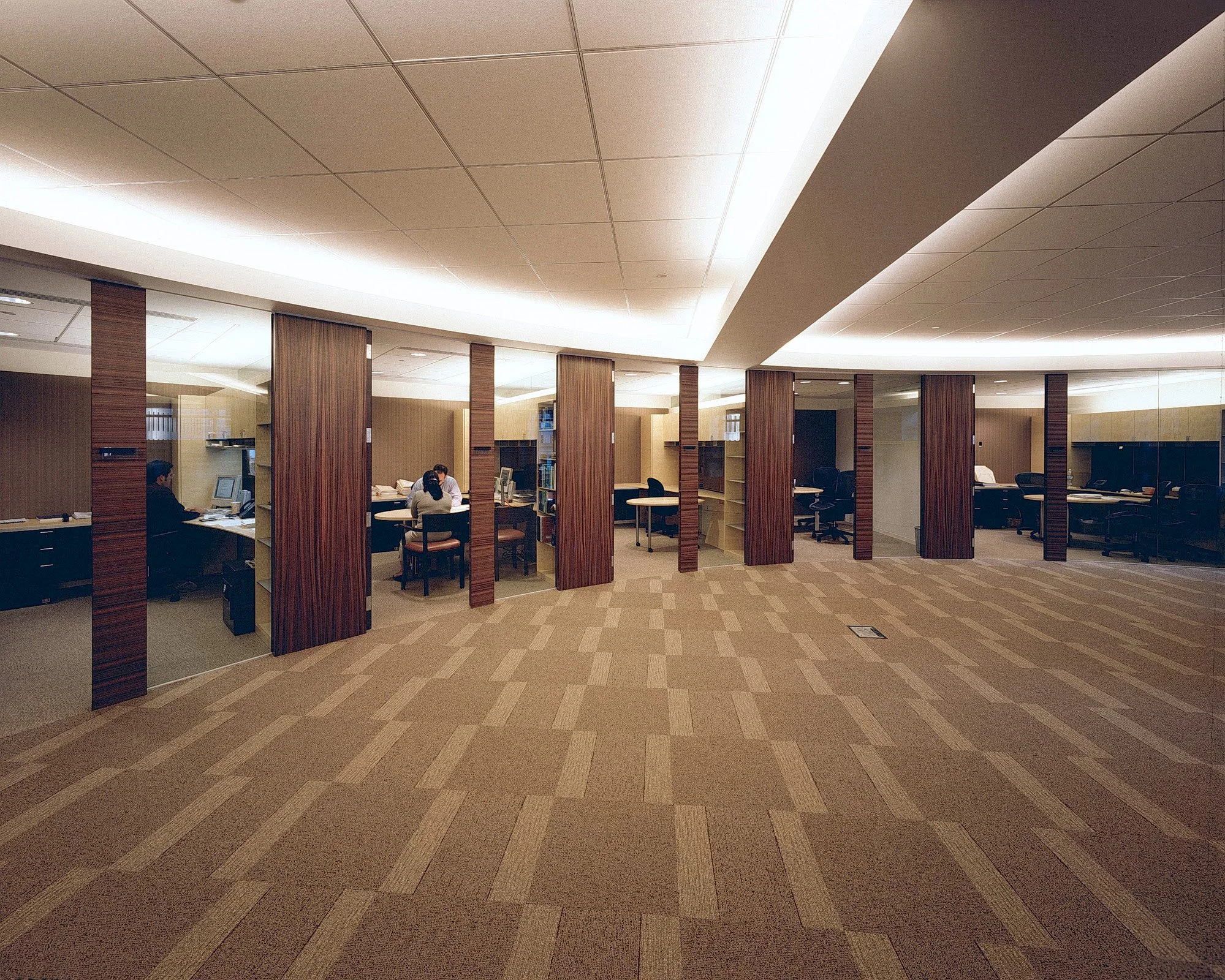 An empty office space with a row of cubicles on either side and a large open area in the center. The cubicles have dark furniture, and some people are working at their desks inside them.