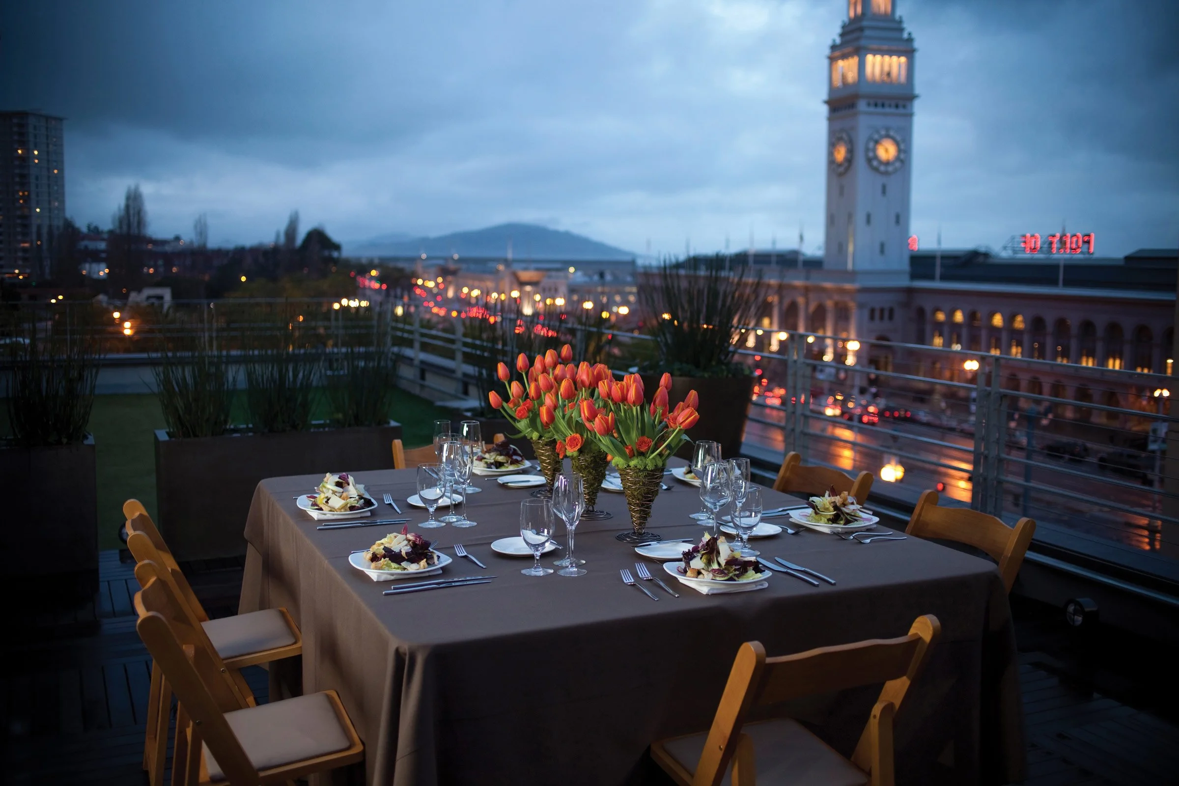 A rooftop dinner table set with plates, utensils, and glasses, featuring a centerpiece of orange tulips, overlooking a cityscape with a clock tower at dusk.