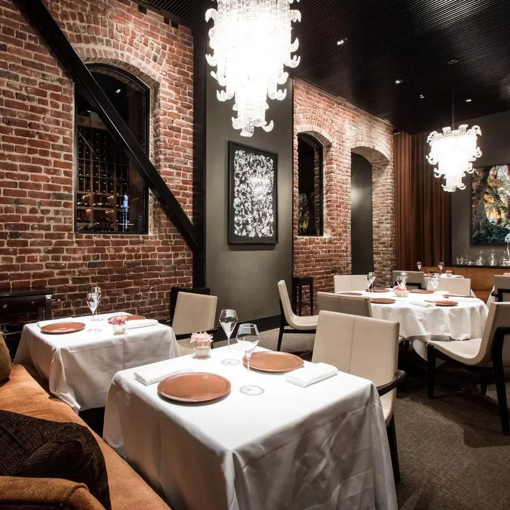 A cozy restaurant dining area with brick walls, white tablecloth-covered tables, and elegant chandeliers hanging from a dark ceiling. The tables are set with napkins, plates, glasses, and small floral centerpieces.