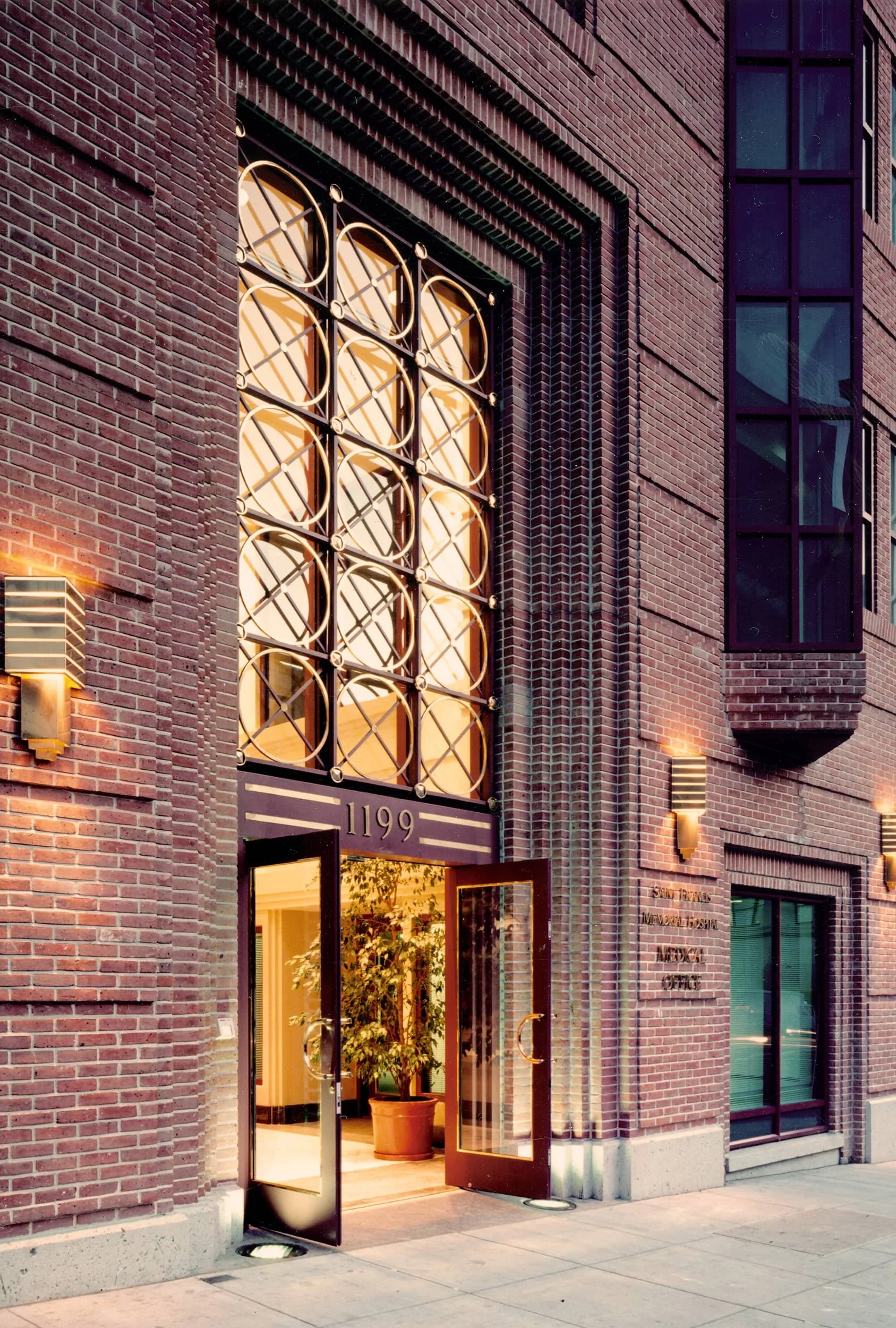 Exterior view of a brick building with the street address 1199 above a glass door entrance, inside a large potted plant is visible.