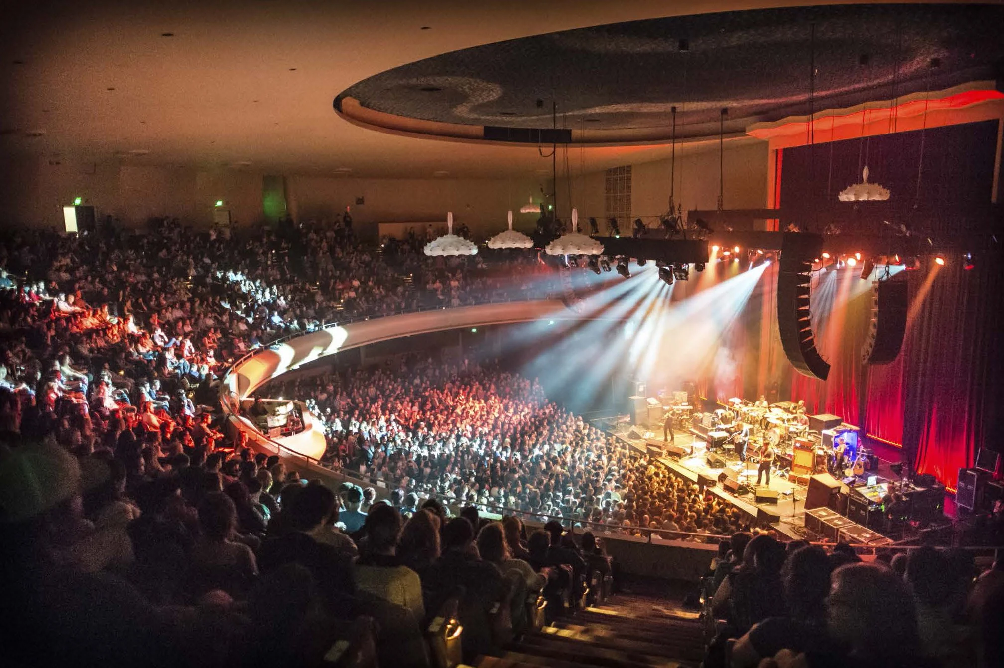Concert hall with a stage illuminated by bright lights, a band performing, and a large audience seated facing the stage.