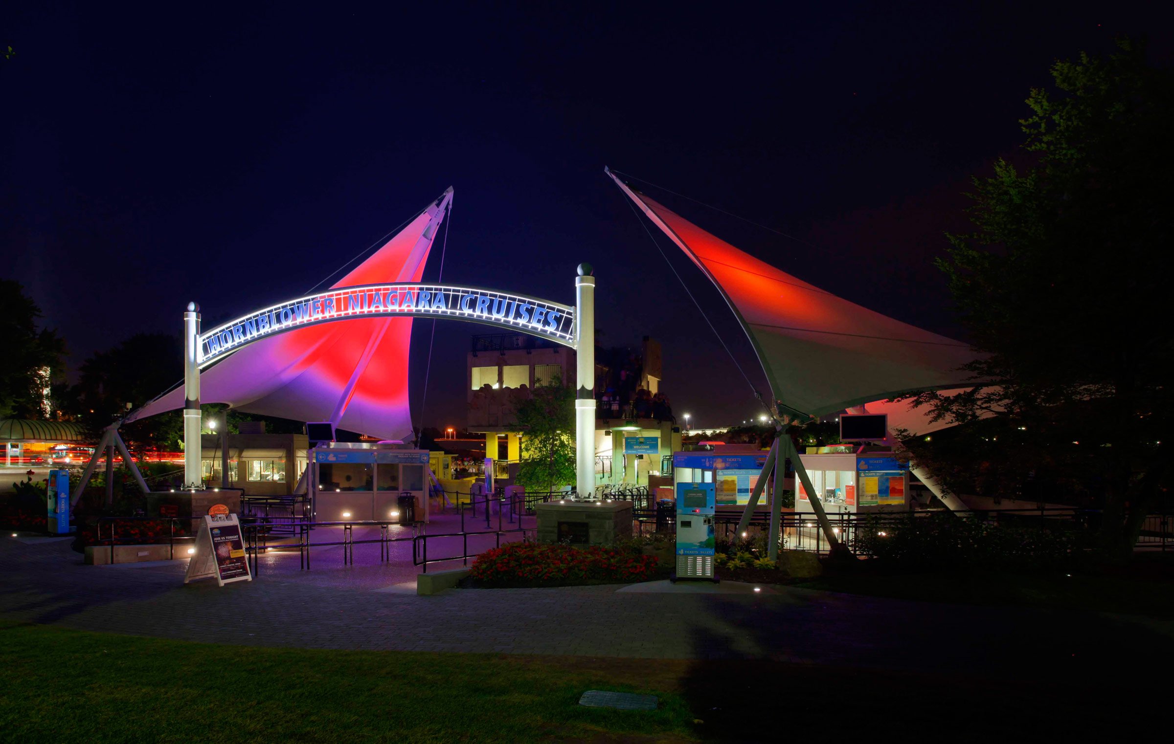 Night view of Hornblower Niagara Cruises with illuminated sails and sign, featuring ticket booths and surrounding trees.