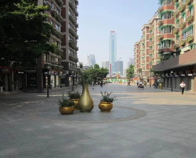 Empty urban plaza with large golden decorative vases and potted plants in the center, surrounded by mid-rise residential buildings, with a city skyline and a tall skyscraper in the background.