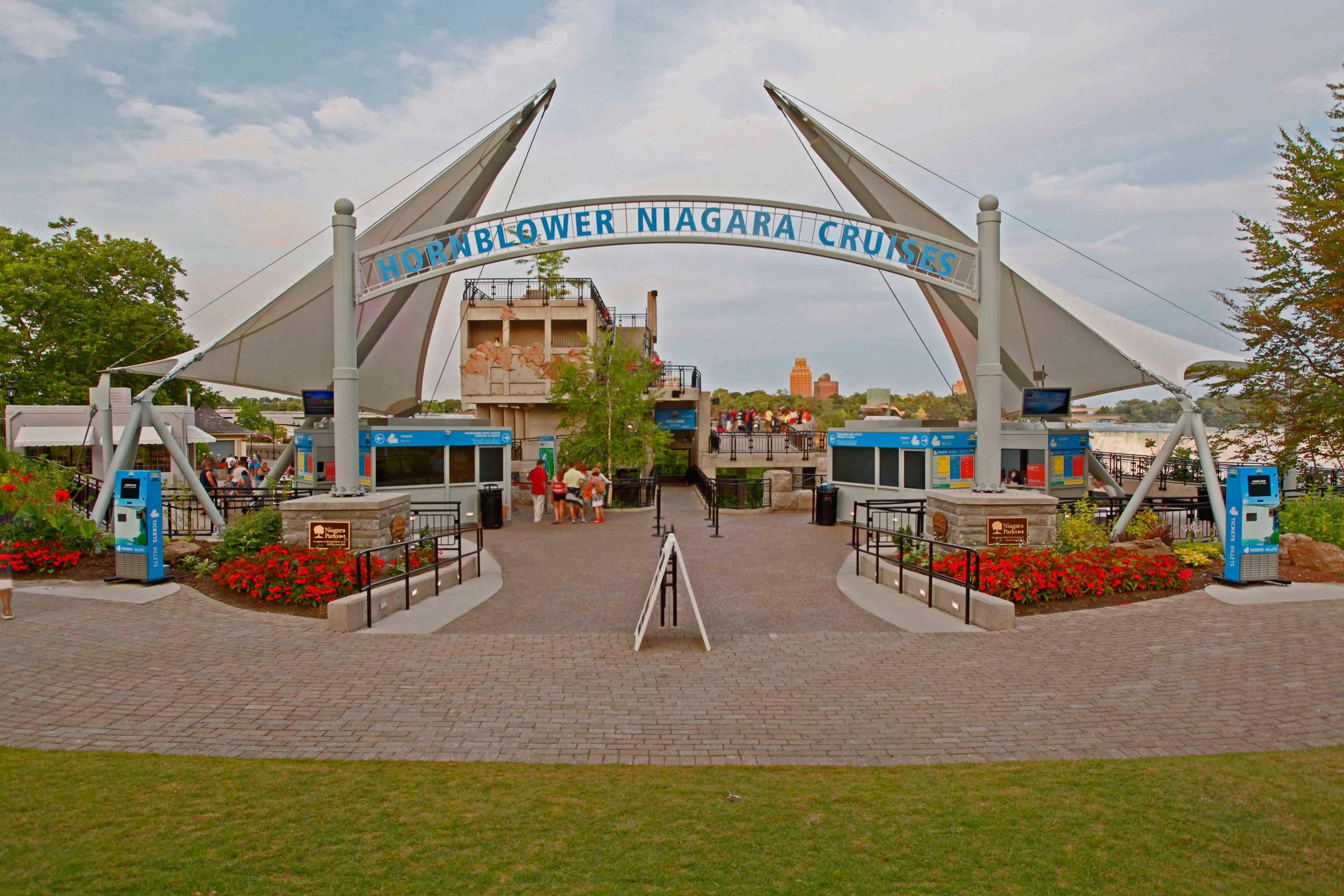 Entrance to Hornblower Niagara Cruises with a large archway sign, canopy structures, visitors, ticket booths, and surrounding flower beds.