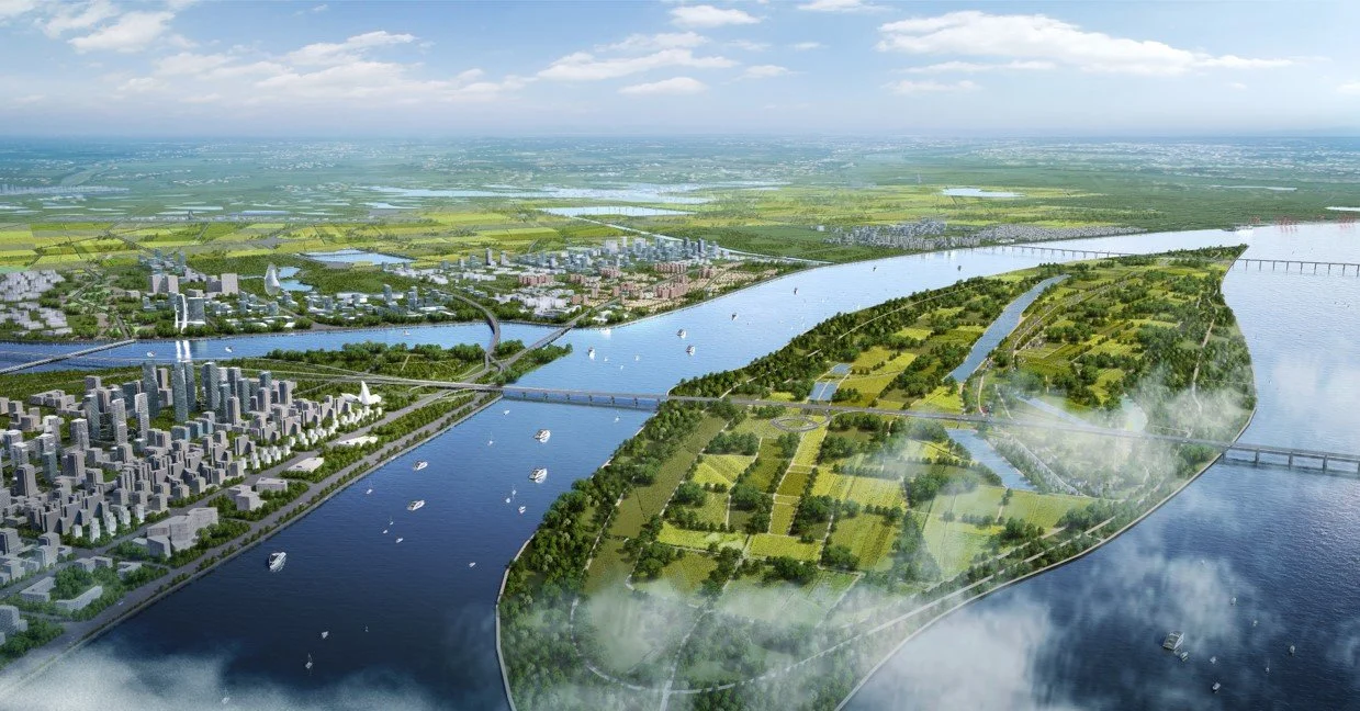 Aerial view of a city and river with boats and bridges, surrounded by parks, green fields, and distant countryside.