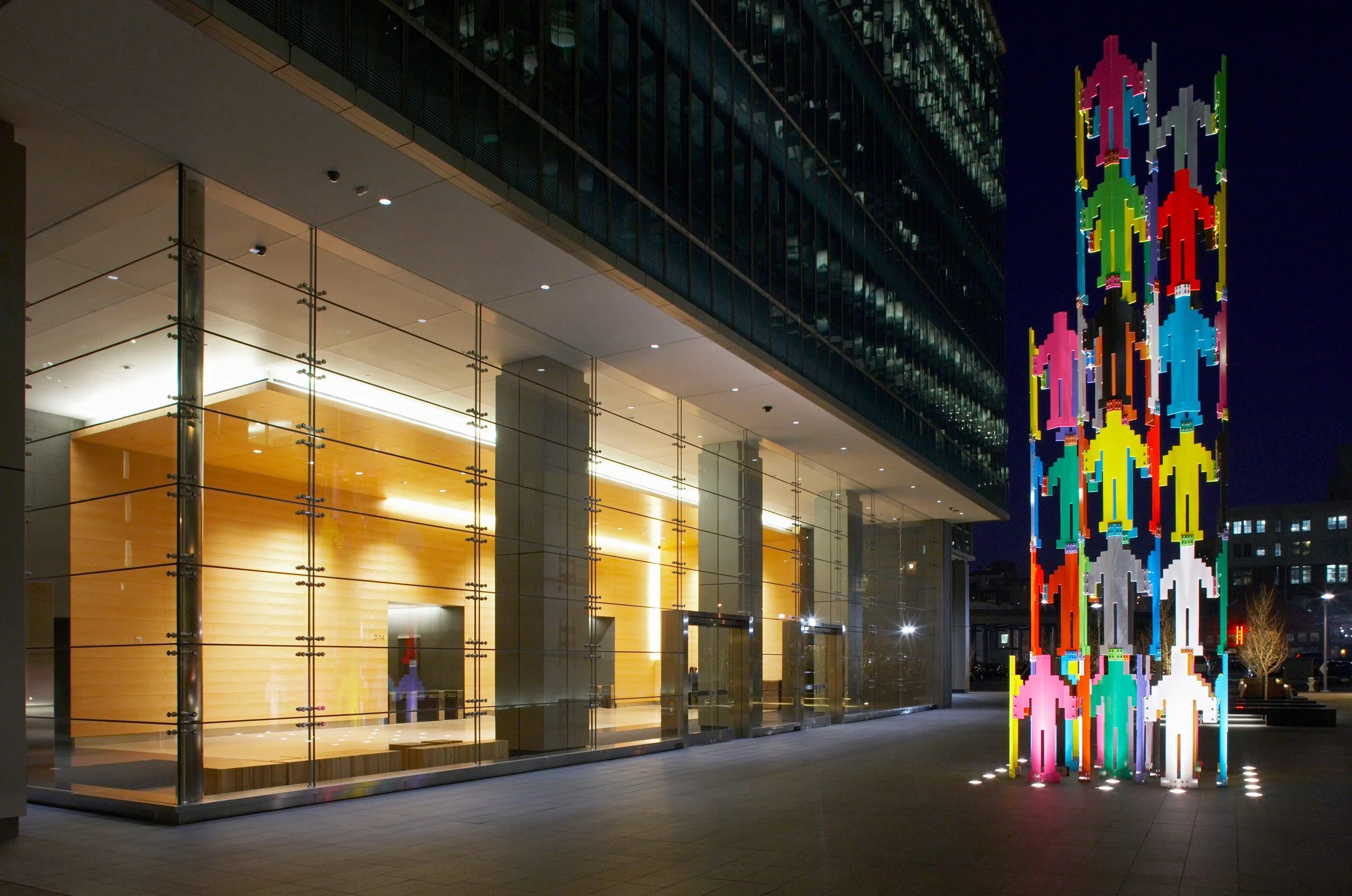 Nighttime view of a modern building with glass walls and warm interior lighting, next to a colorful illuminated public art sculpture of stylized human figures in bright colors arranged in a column.
