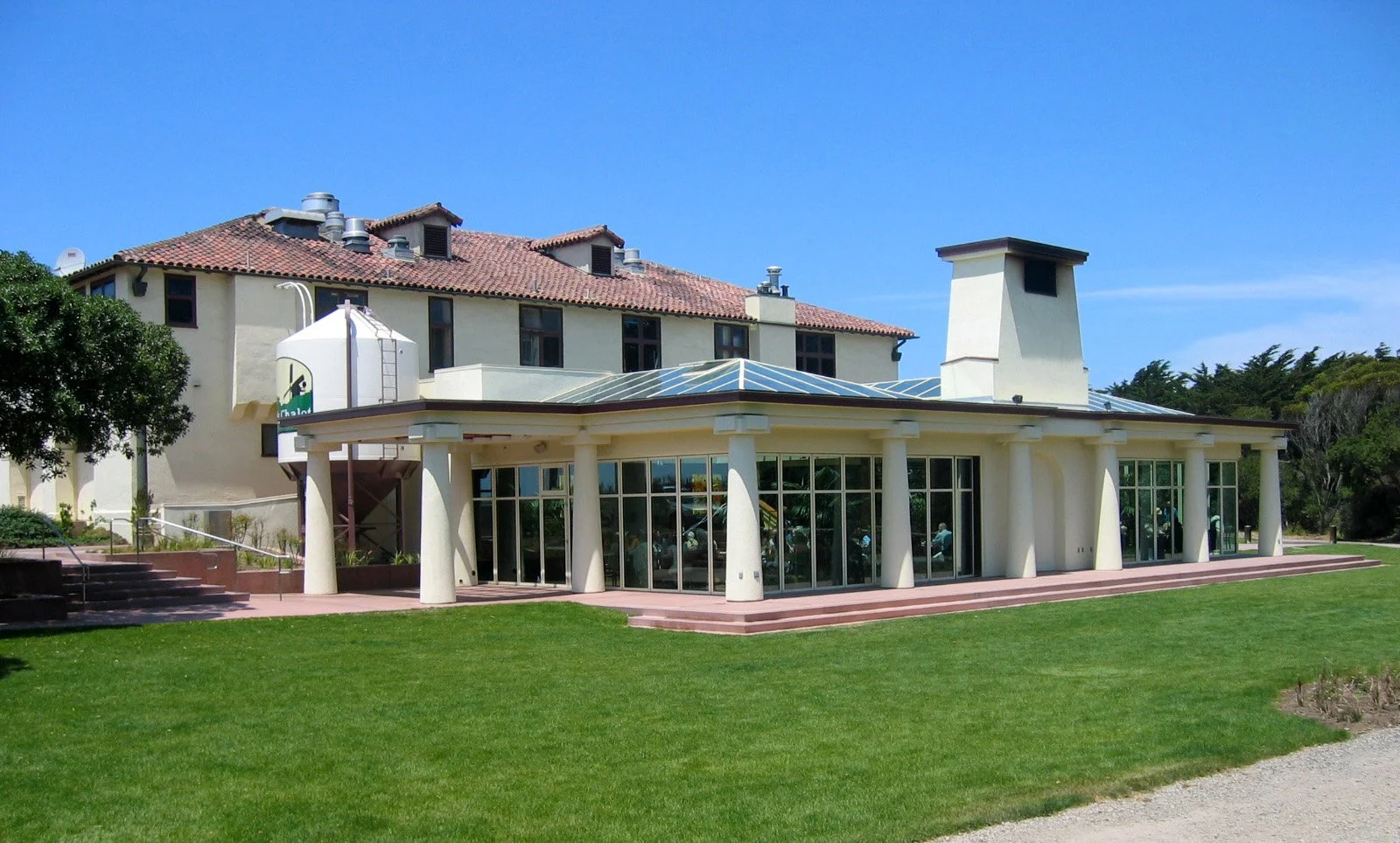 A modern building with large glass windows and a unique architectural design, featuring a white facade, cylindrical columns, and a sloped roof, surrounded by a well-maintained lawn under a clear blue sky.