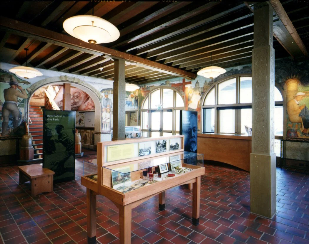 Interior of a museum or historical exhibit space with display cases, informational panels, mural paintings, and large arched windows.