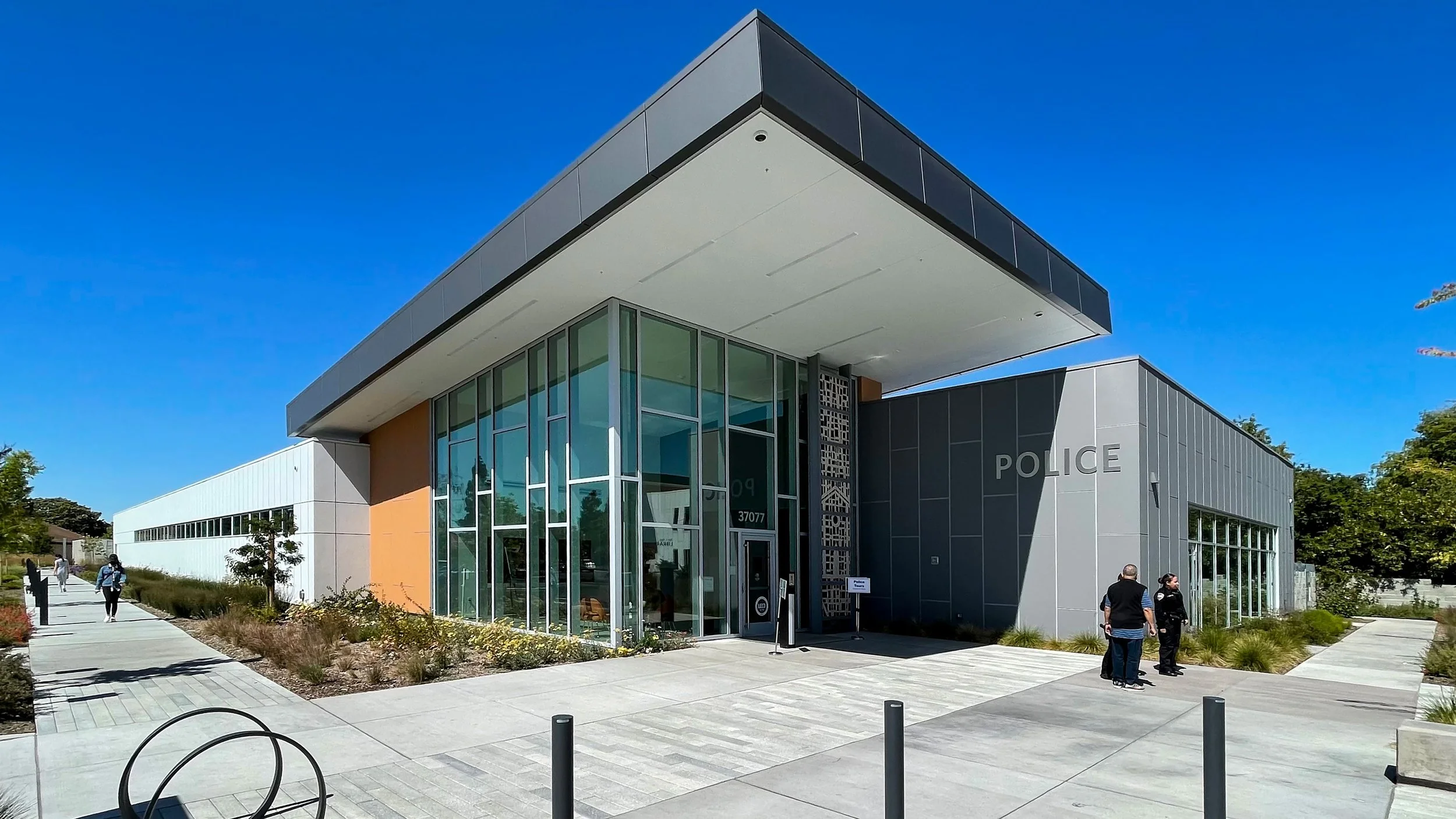 Modern police station with glass entrance and gray exterior walls, surrounded by landscaped bushes and trees under a clear blue sky, with people walking and talking outside.