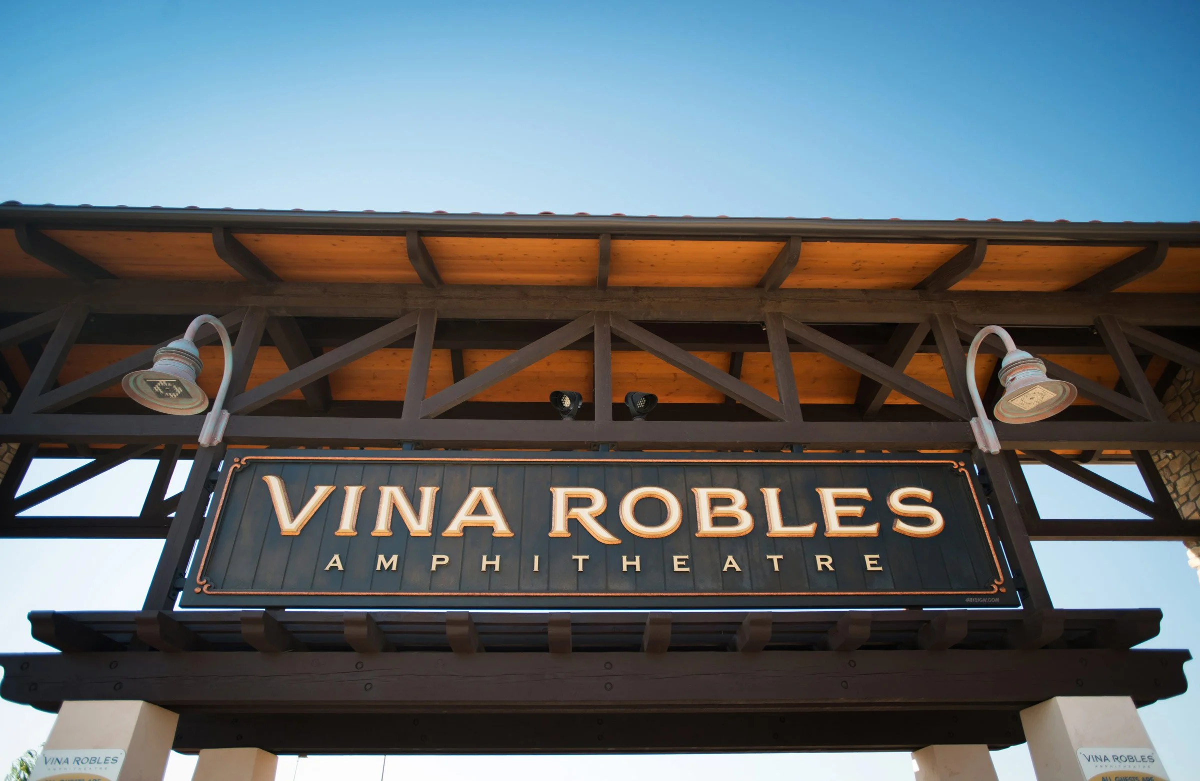 Sign for Vina Robles Amphitheatre mounted on a wooden structure with spotlights and a clear blue sky in the background.