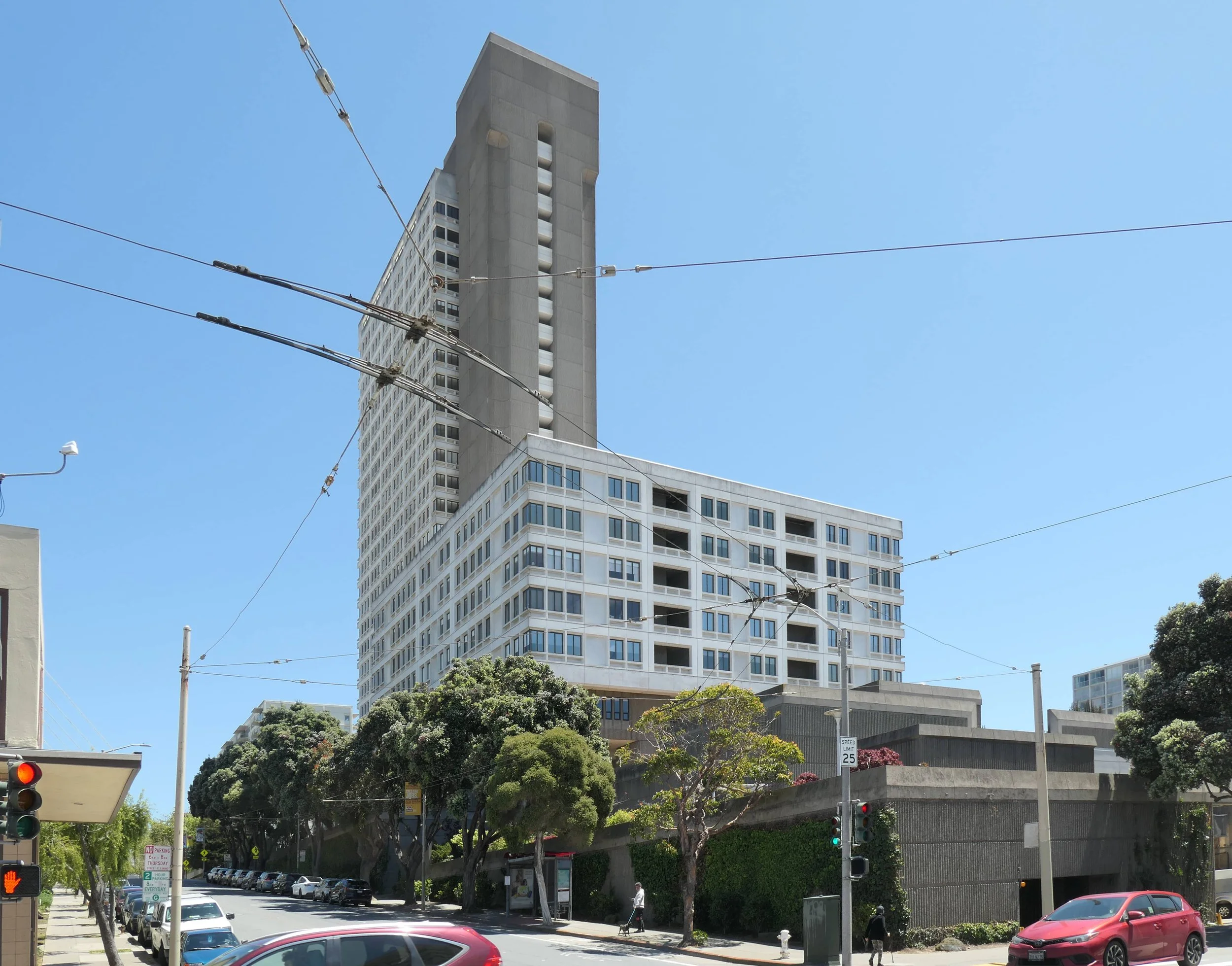 A modern high-rise building with a unique design, featuring a tall, rectangular tower connected to a shorter, wide section with multiple windows, located on a city street with trees, cars, and traffic lights.