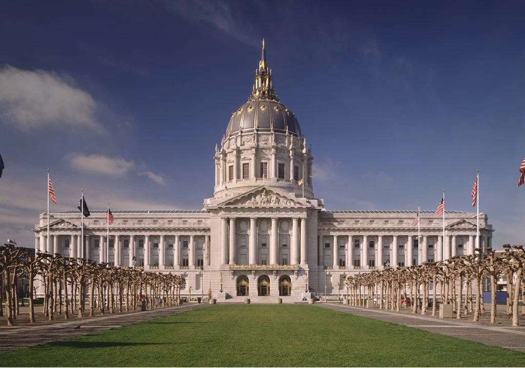 The San Francisco City Hall, a grand historic building with a large central dome, surrounded by manicured trees and American flags, under a partly cloudy sky.