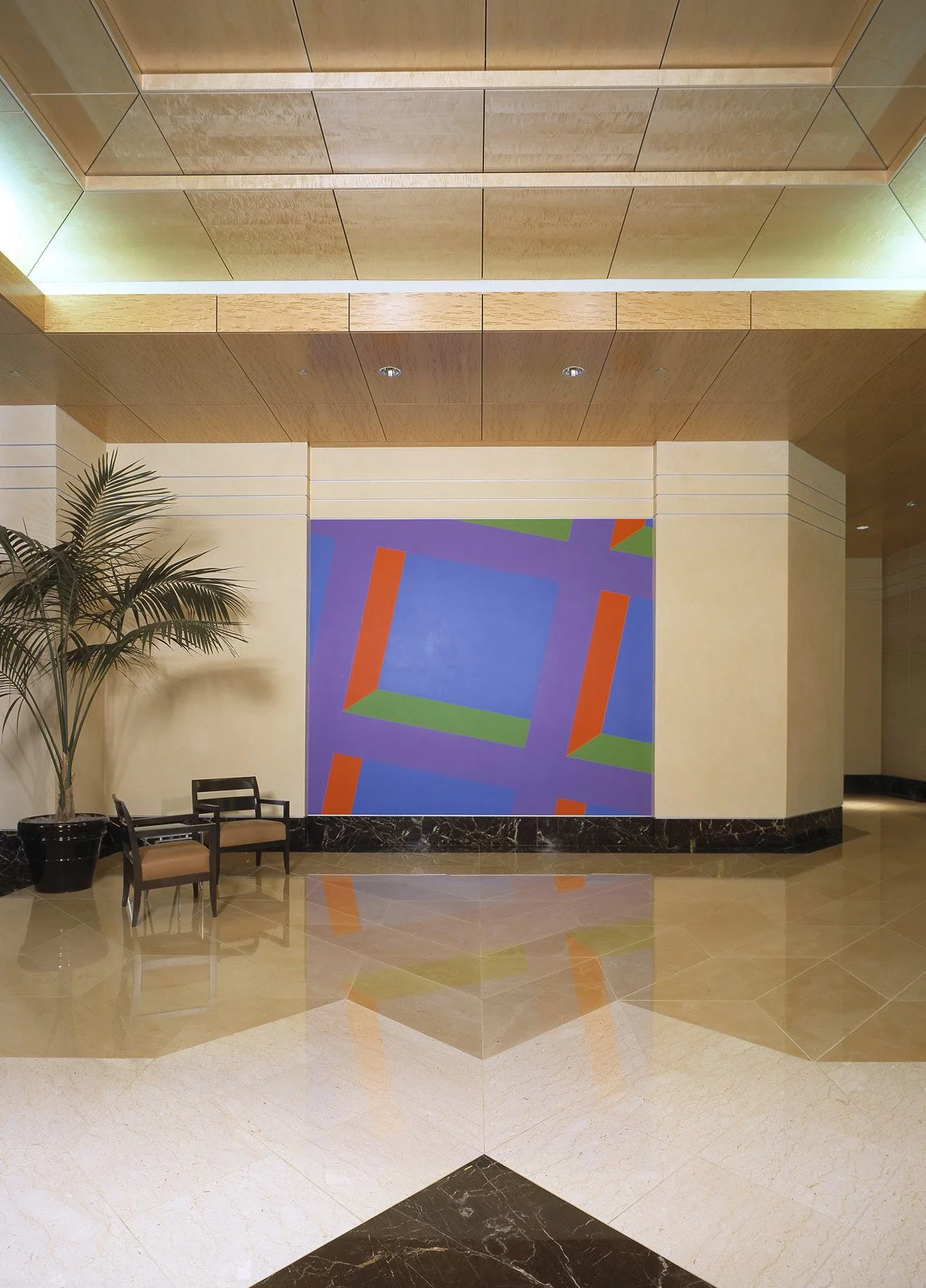 Modern hotel lobby with abstract wall art, a potted plant, and seating area with two chairs and a small table.
