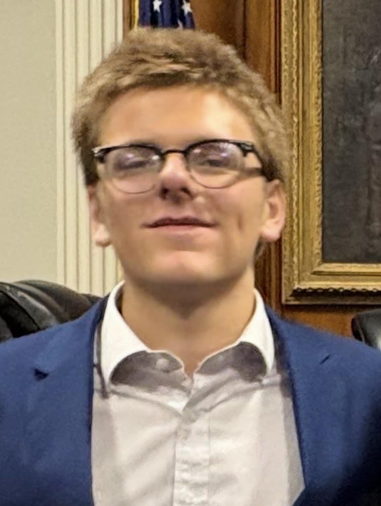 Young man with glasses, wearing a white shirt and a blue blazer, smiling in front of a framed picture and American flag.