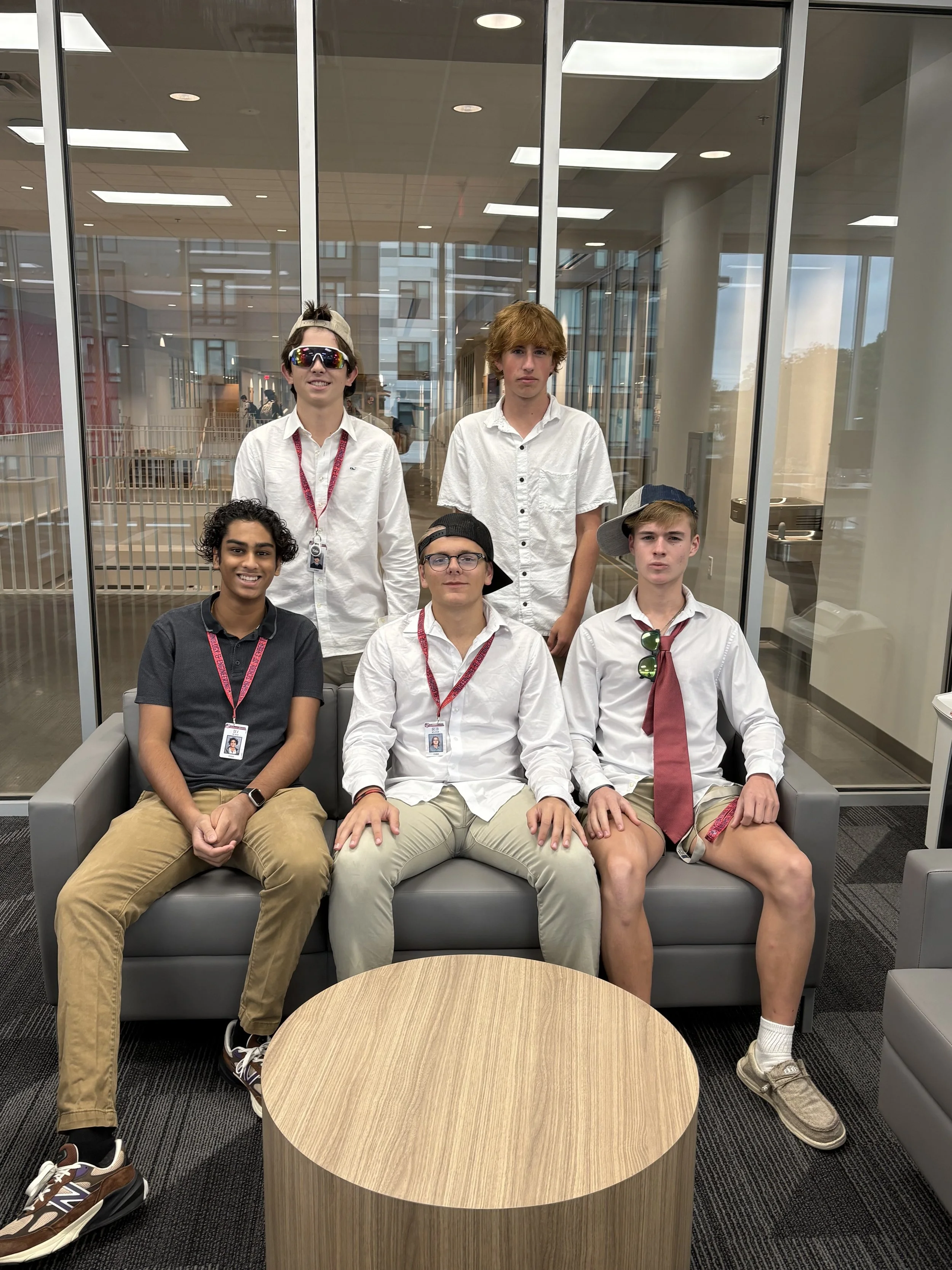Six teenage boys wearing casual and semi-casual clothing, some with accessories like sunglasses and hats, sitting and standing in an indoor lounge area with glass windows behind them.