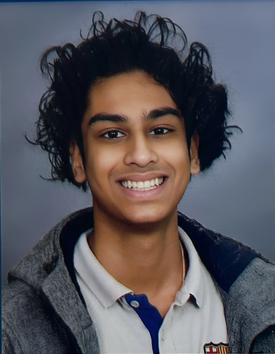 Young man with curly black hair, smiling, wearing a white shirt with a buttoned collar and a dark jacket, against a gray background.