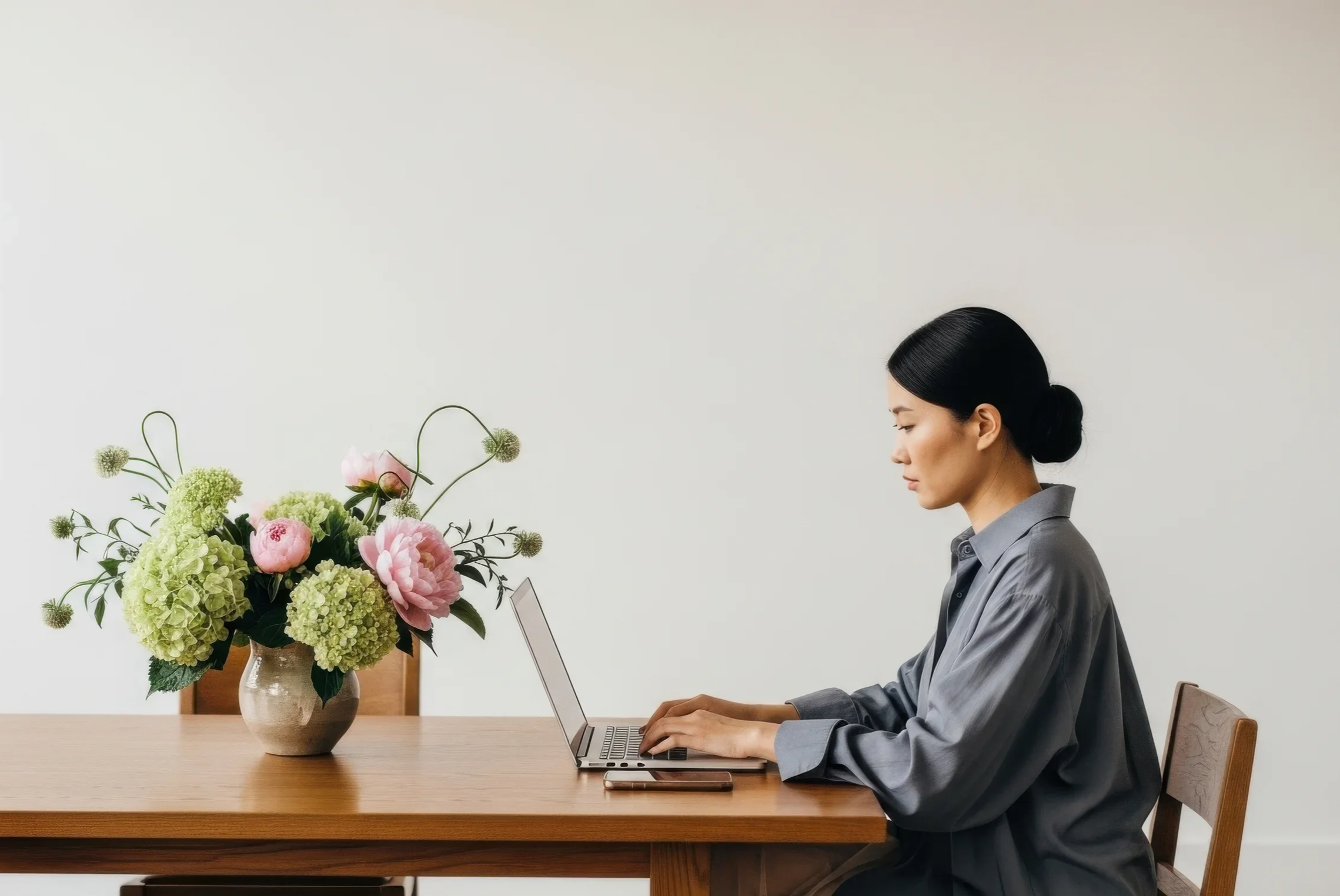 Woman with black hair tied in a bun sitting at a wooden table, working on a laptop, with a vase of pink and green flowers on the table.