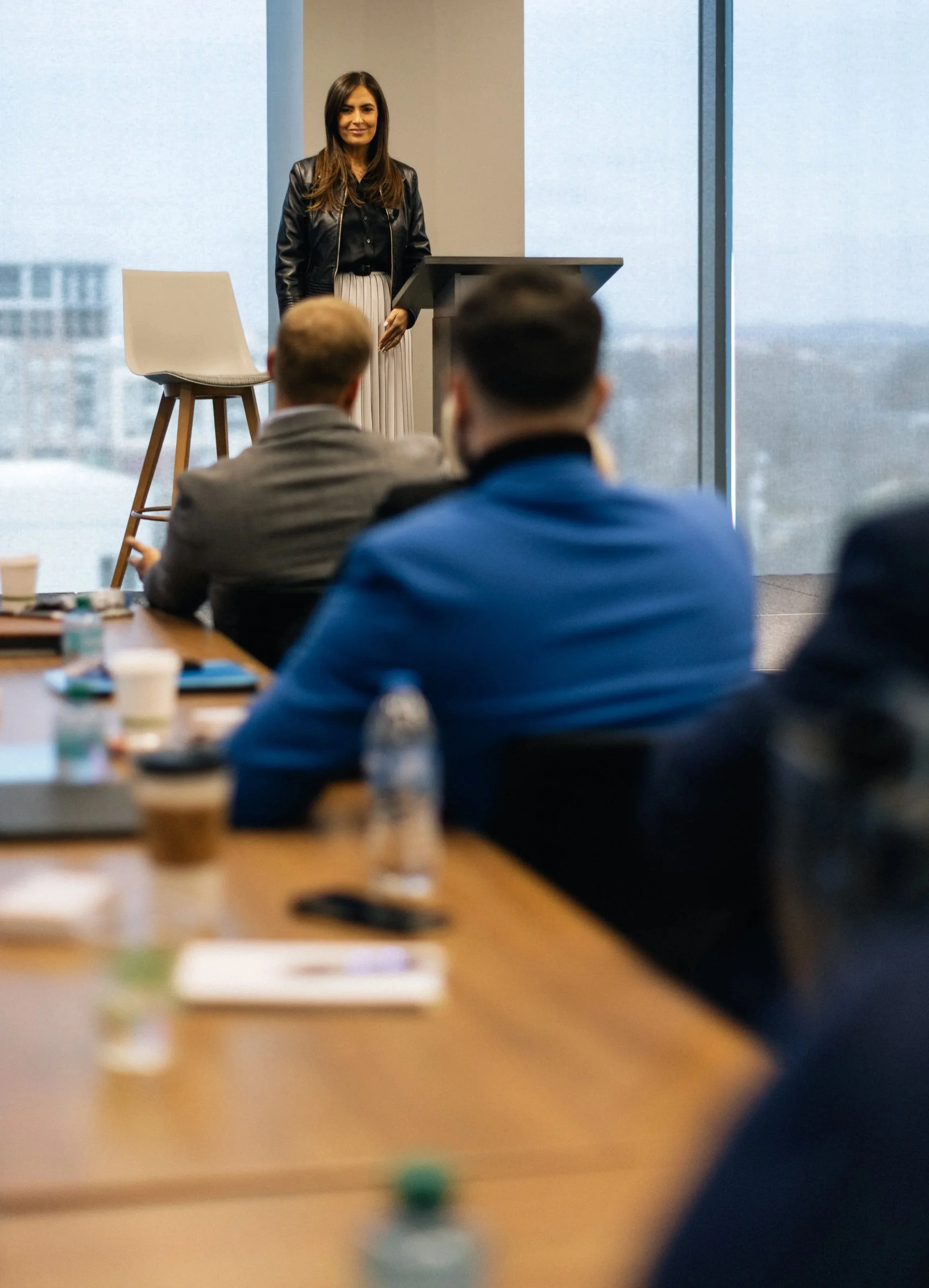 A woman giving a presentation in a modern office conference room with attendees seated at a table, and large windows showing a cityscape in the background.