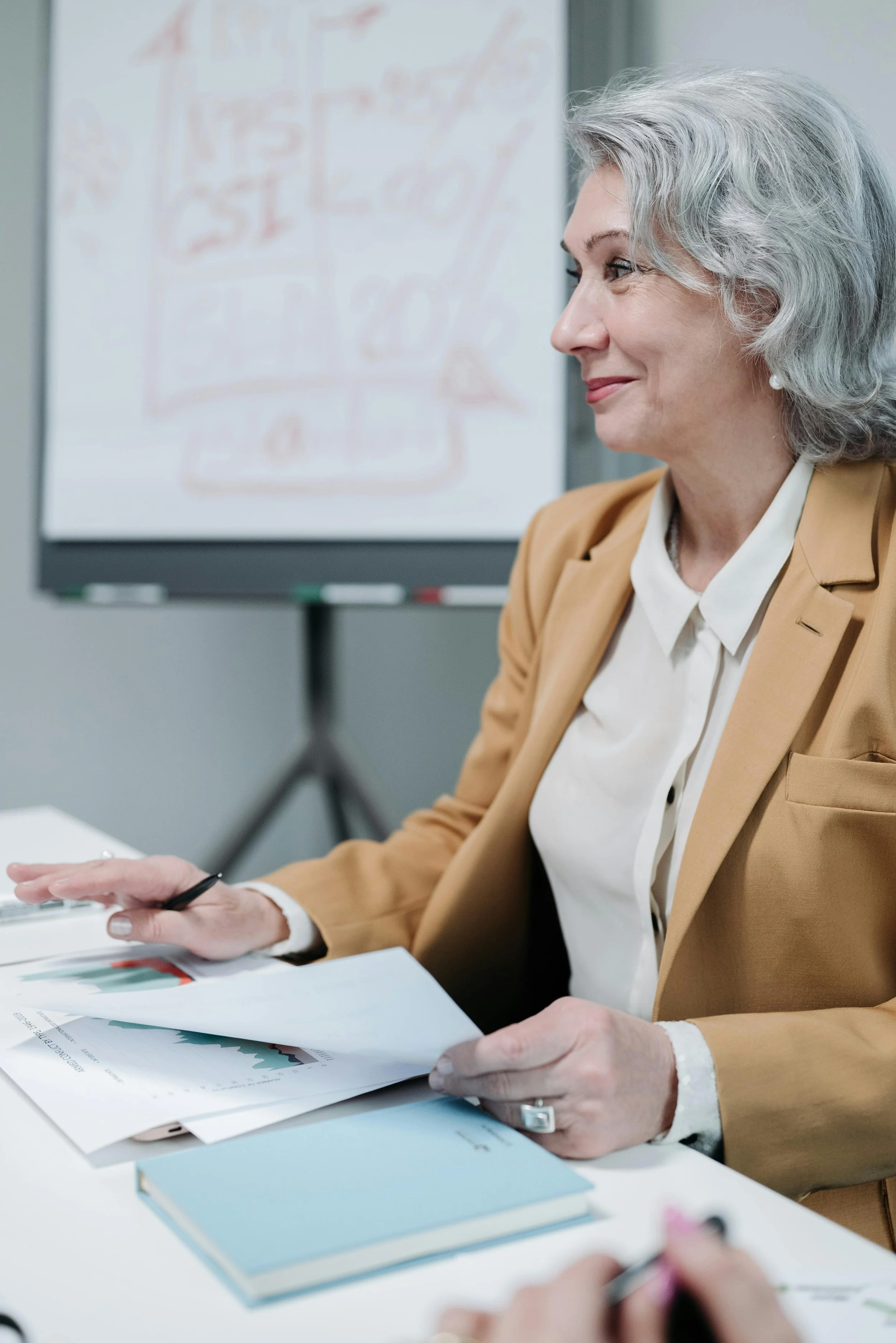 A professional woman with gray hair in a beige blazer and white blouse is sitting at a desk, examining and discussing printed documents with graphs during a meeting. There's a whiteboard with red writing in the background.