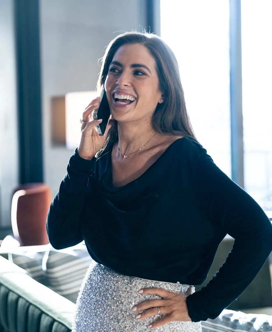 A woman with long brown hair, wearing a black top and a silver sequined skirt, is smiling and talking on a cell phone indoors near a window.