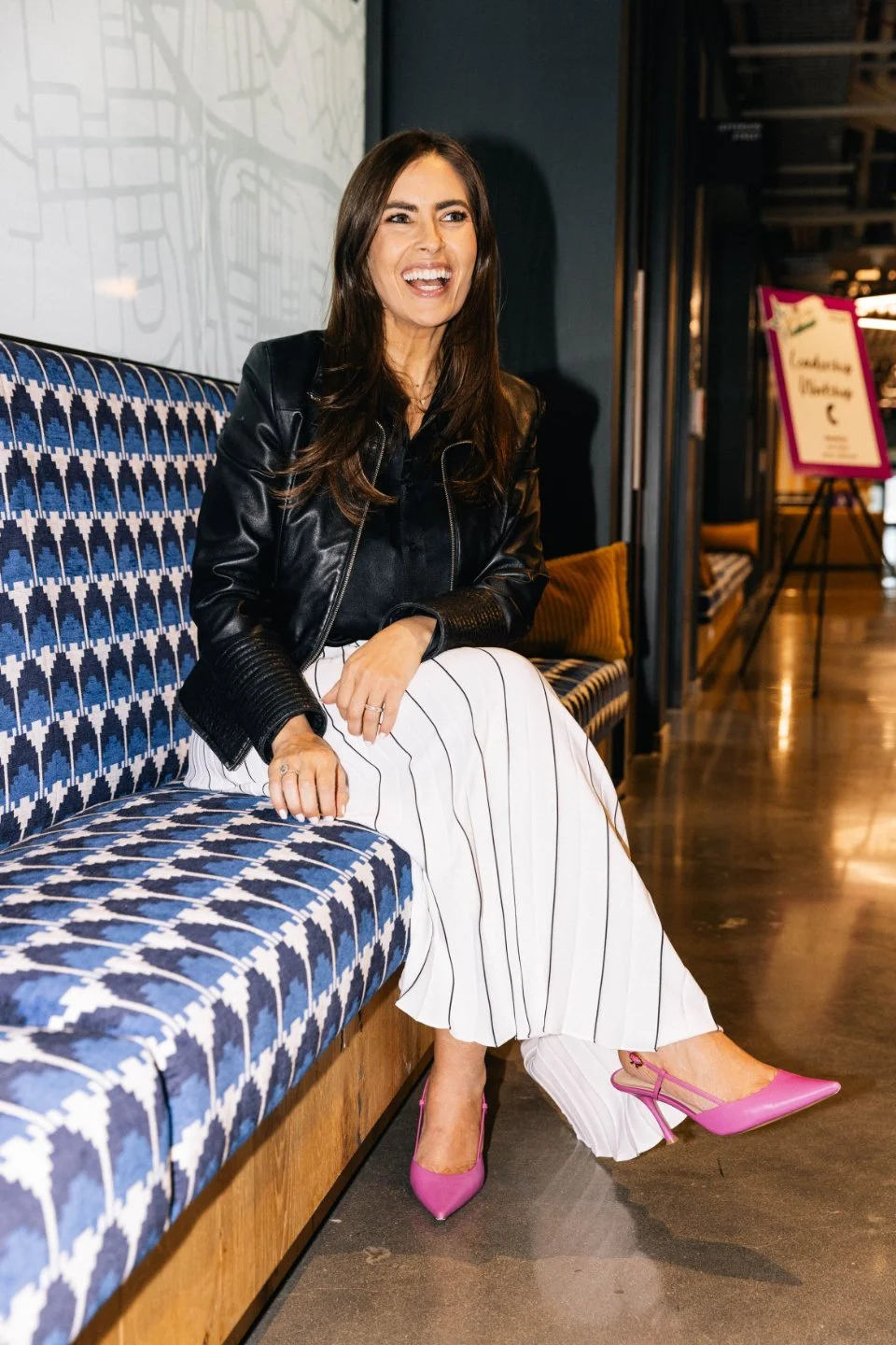 A woman with long brown hair sitting on a patterned blue and white bench, smiling, wearing a black leather jacket, a white pinstripe skirt, and pink high heels.