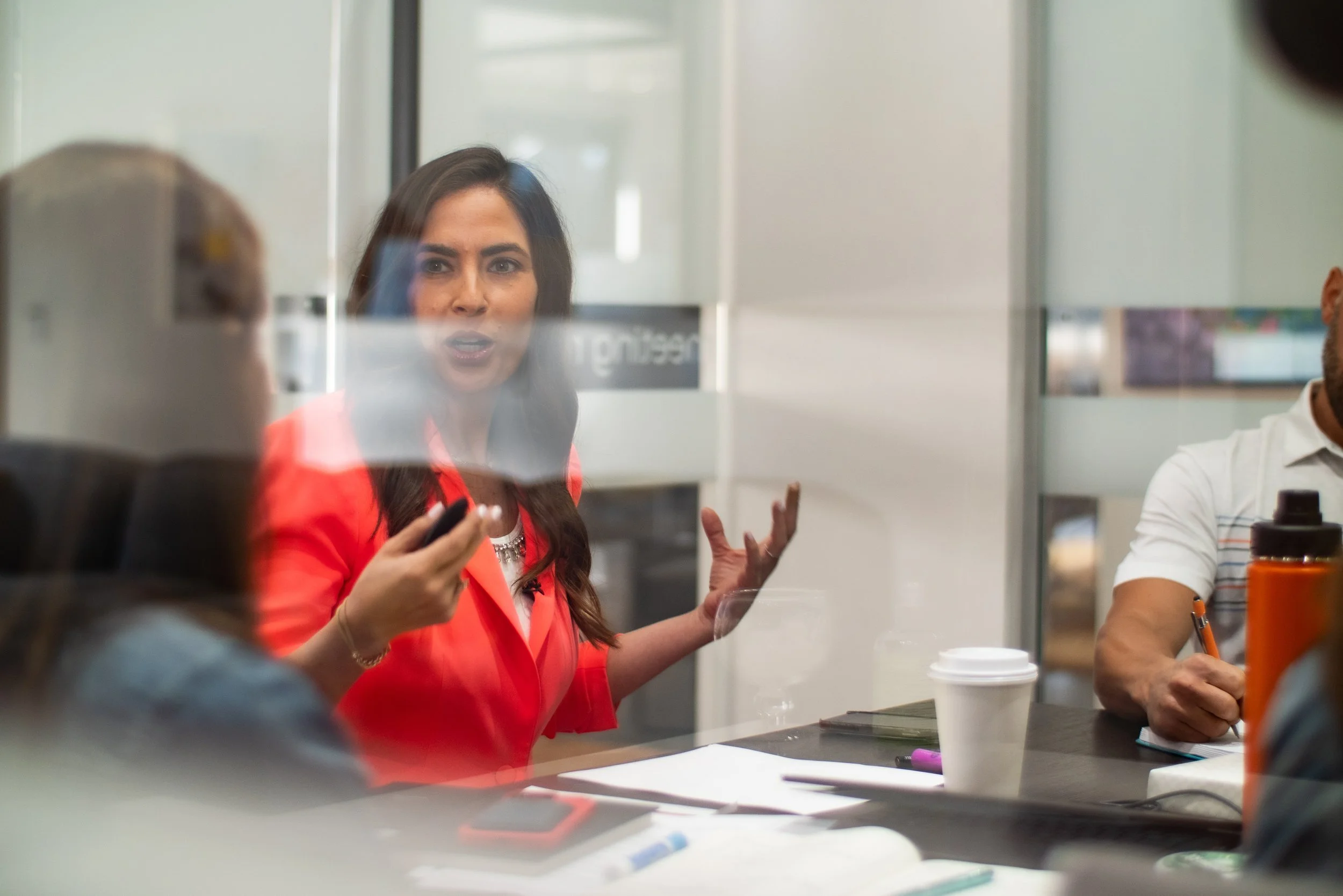 A woman with long dark hair wearing a red jacket is sitting at a conference table, speaking passionately and gesturing with her hands, while holding a remote or phone. There are papers, a notebook, a pen, and a coffee cup on the table. Other people are partially visible around her.