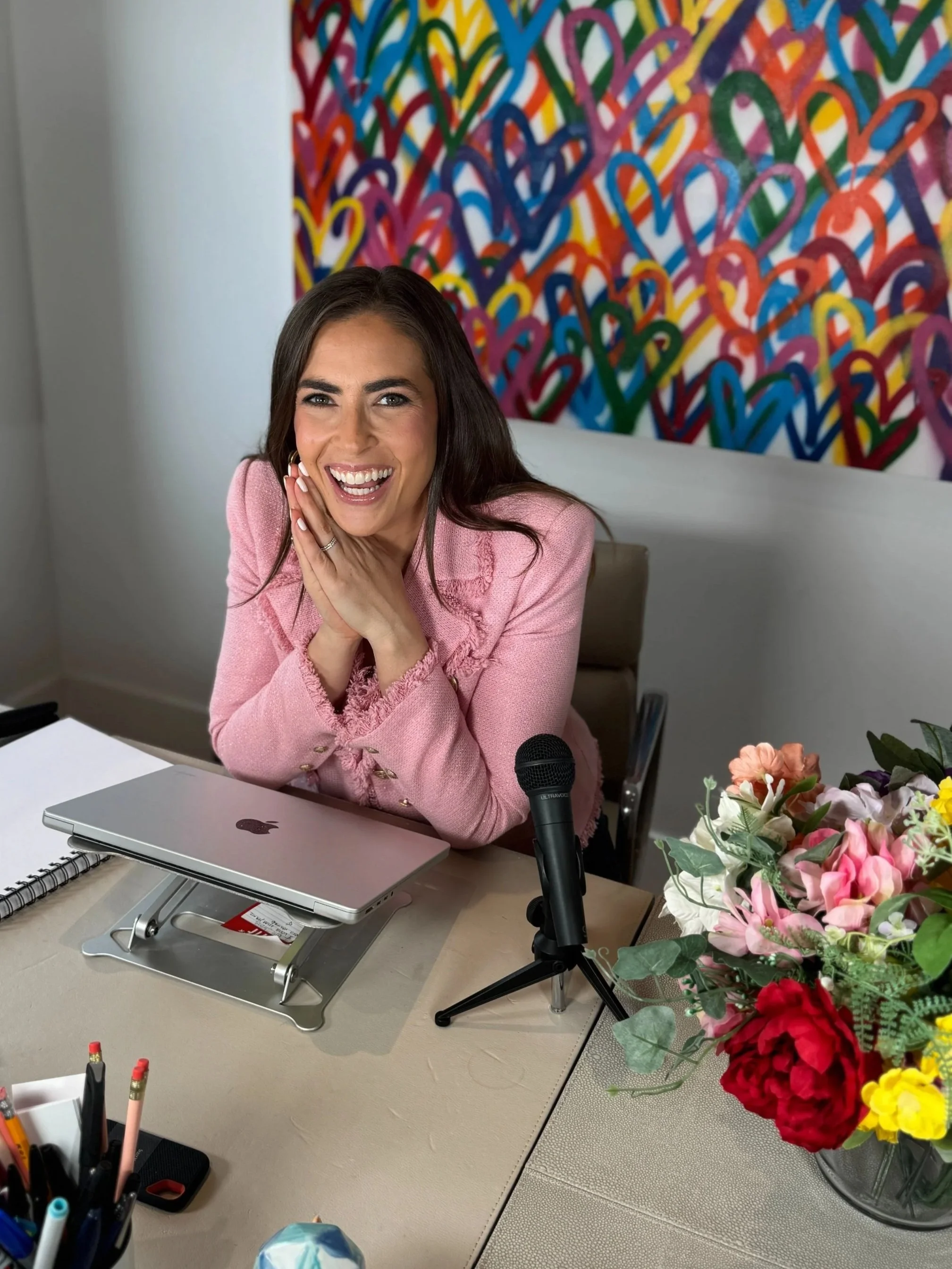 A woman in a pink blazer sitting at a desk with a laptop, microphone, flowers, and colorful heart artwork on the wall behind her, smiling and looking at the camera.