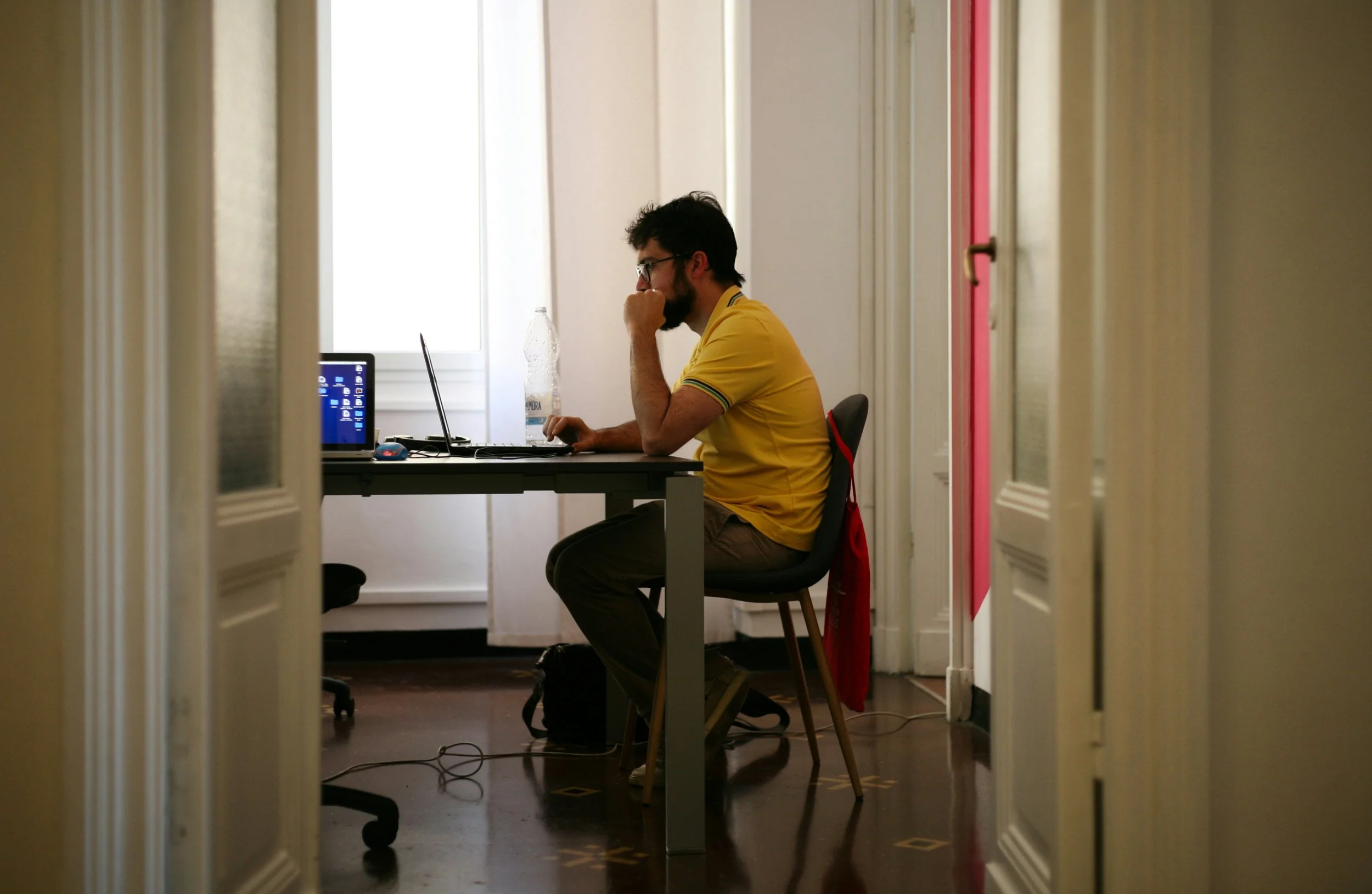 A man wearing a yellow shirt sitting at a desk working on a laptop in a small office.