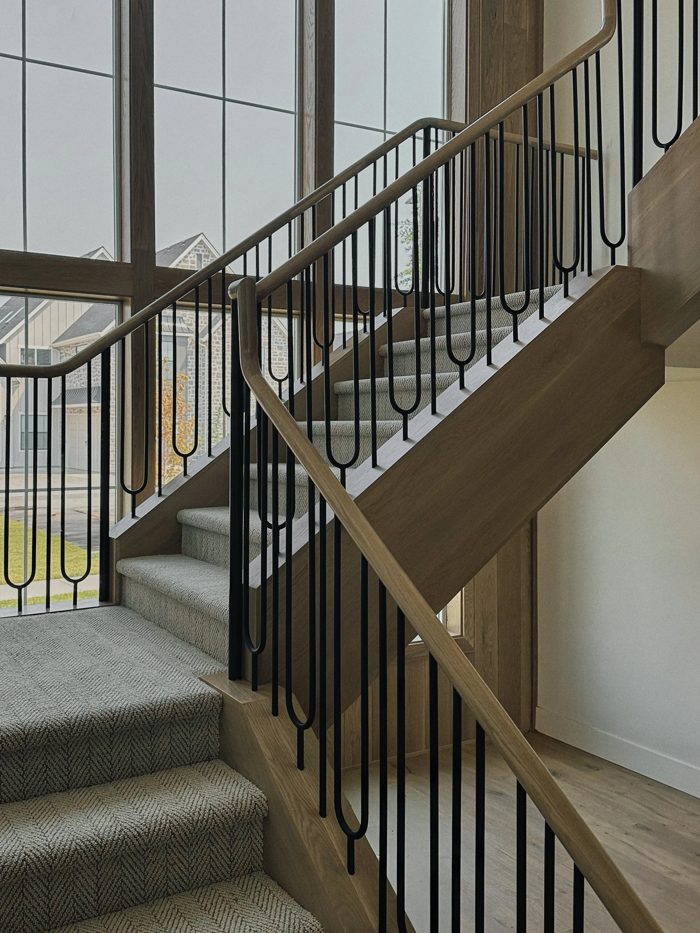 Interior view of a staircase with wooden handrails and black metal balusters, carpeted steps, and large windows letting in natural light.