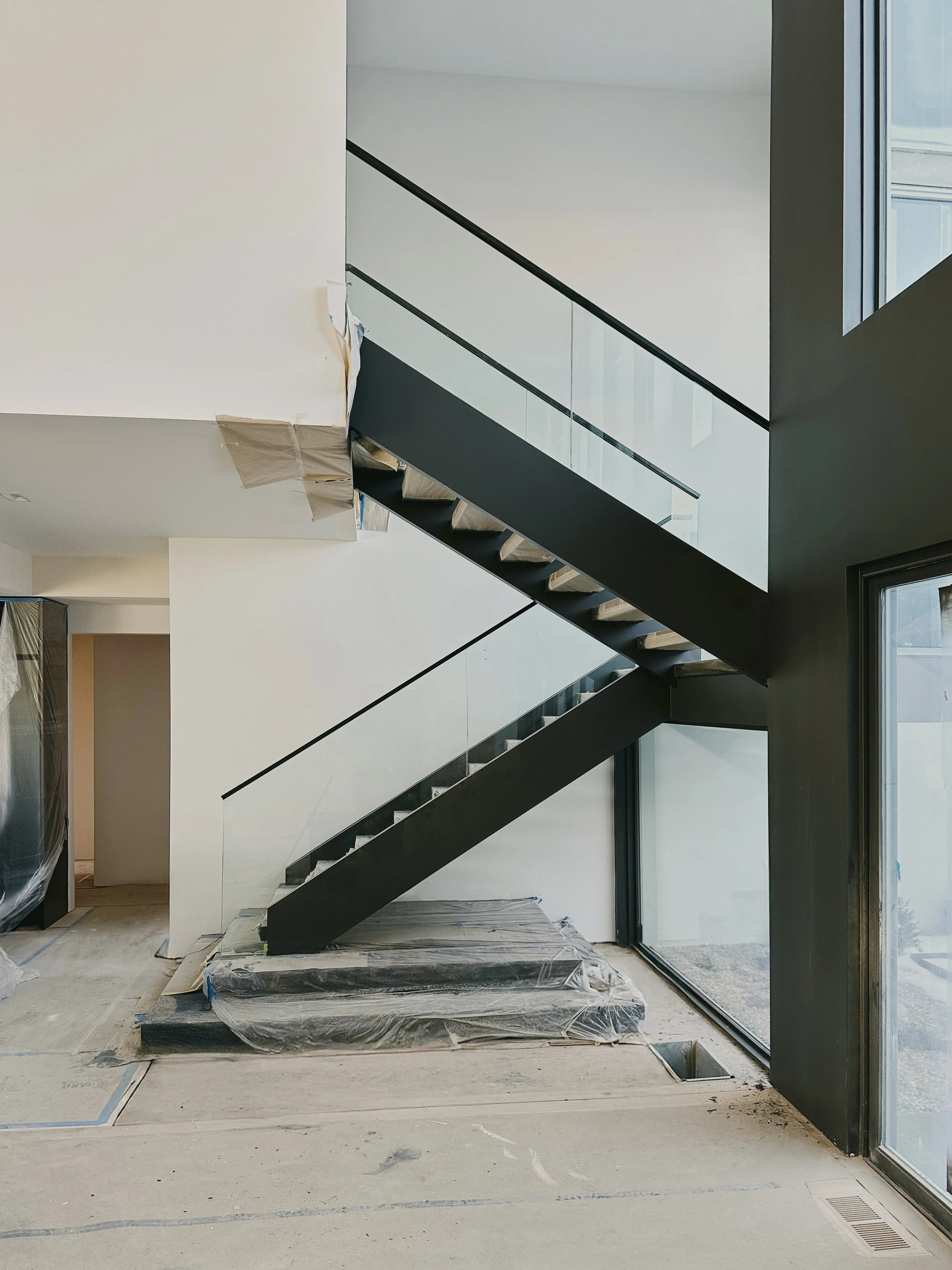Interior of a modern home under construction, featuring a black staircase with glass railings and large windows.