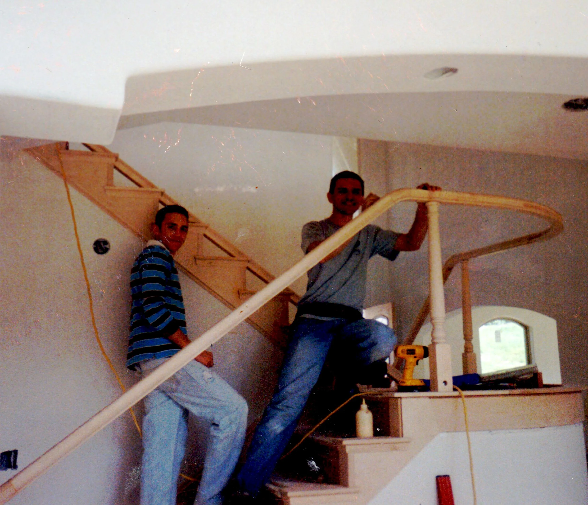 Two men are installing a wooden staircase in a house, one is standing on the lower step and the other is on the upper part of the staircase.