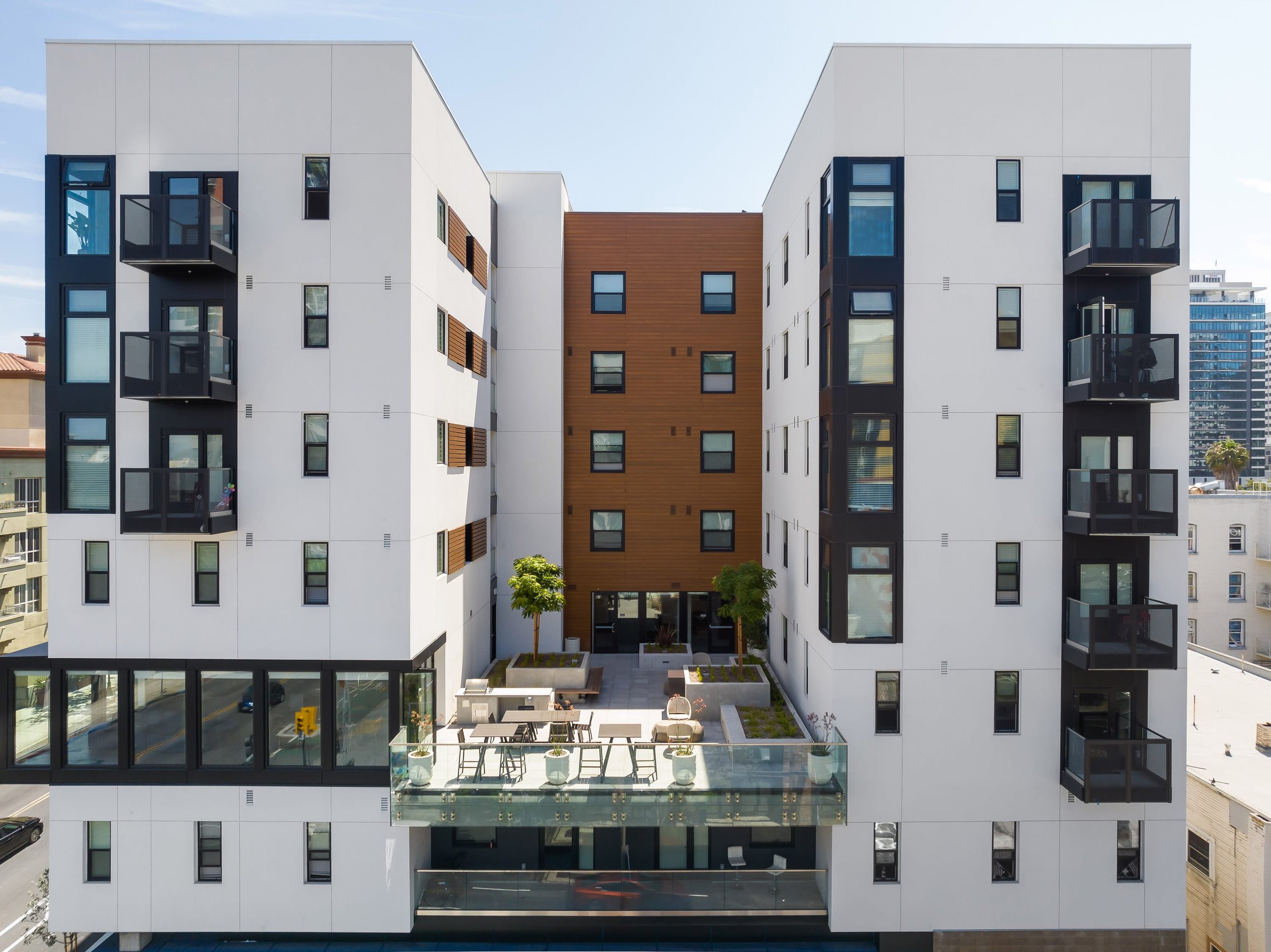 The Helm: a modern multi-story apartment building with white and wooden exterior, glass balcony railings, and a rooftop terrace with seating, trees, and planters.