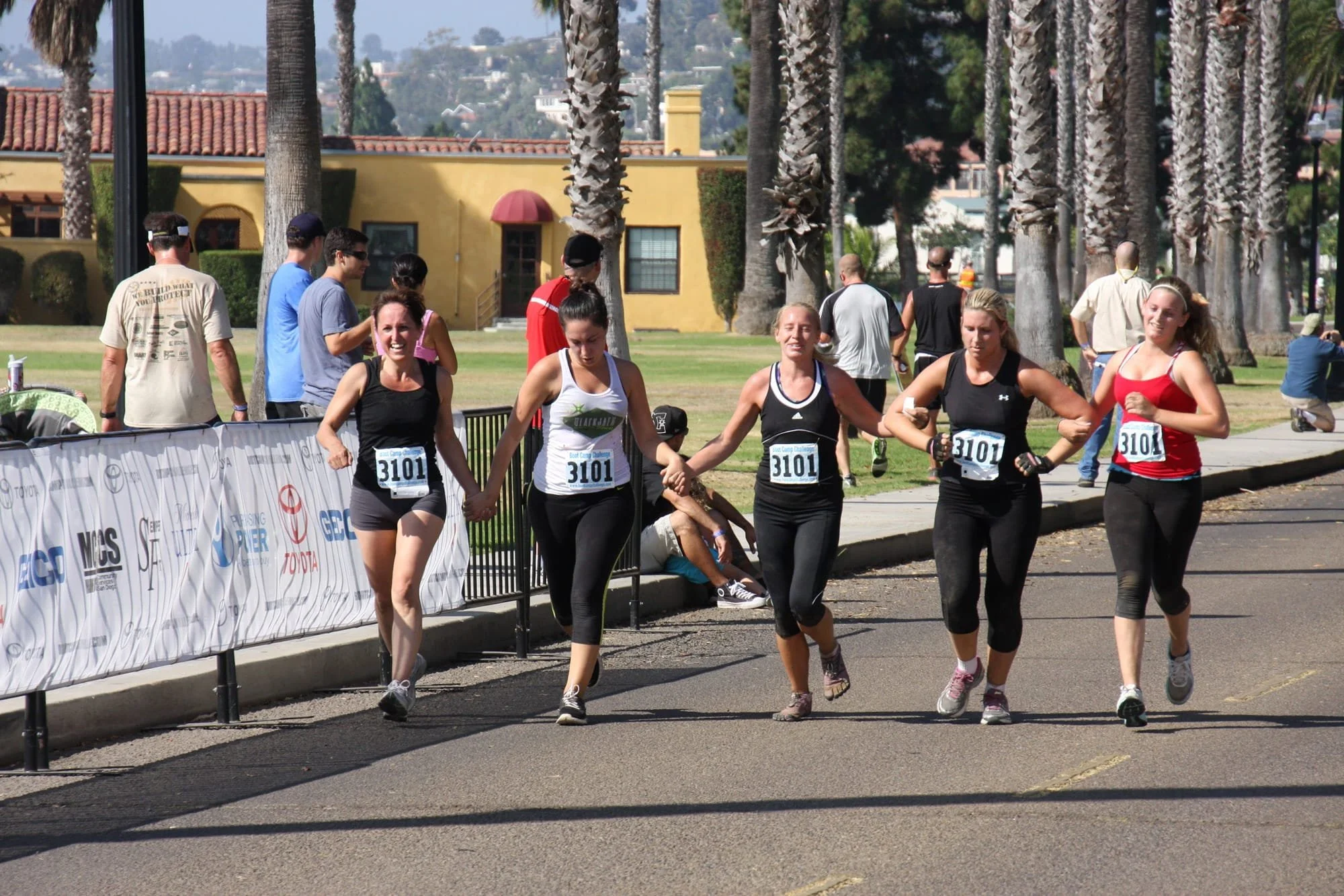 Five women running in a race, holding hands and crossing the finish line outdoors on a sunny day.