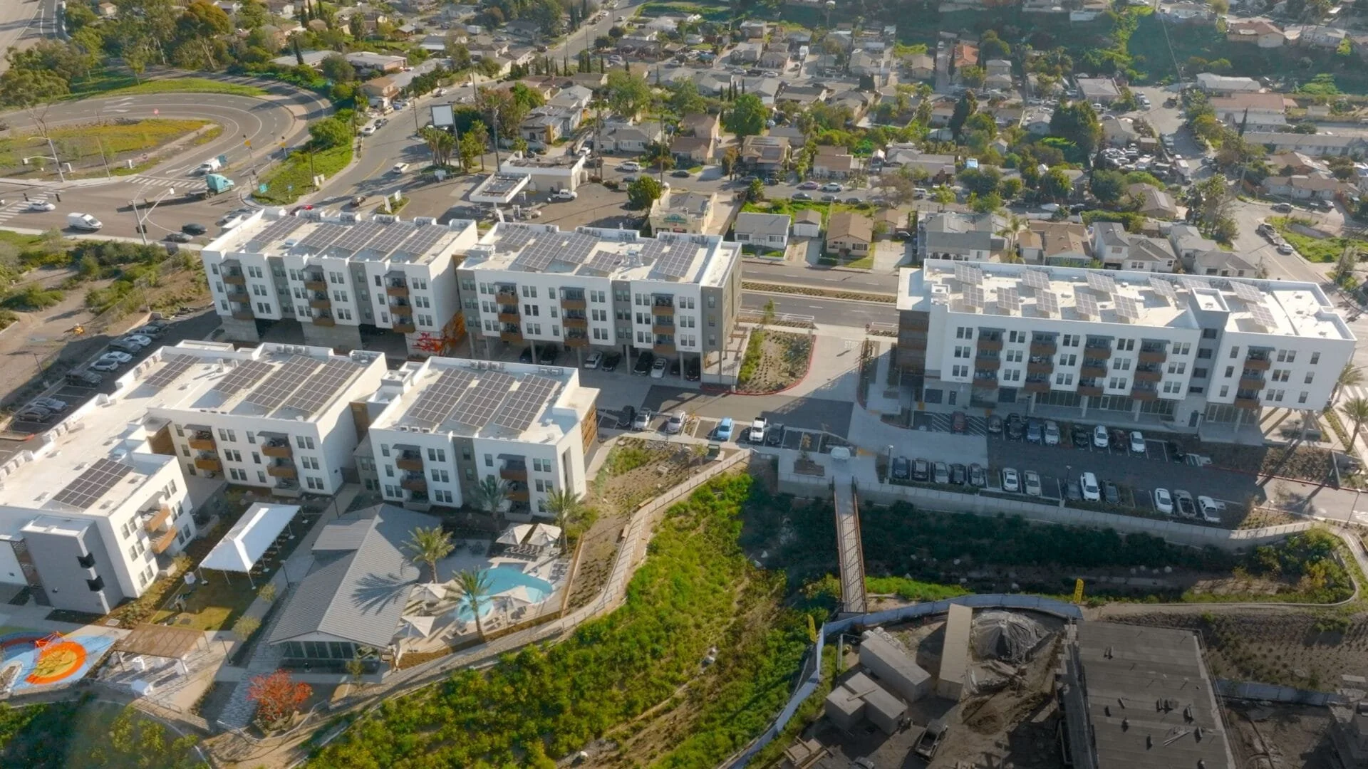 Aerial view of multi-unit residential buildings with solar panels on the roofs, parking lots, a swimming pool with umbrellas, and surrounding streets and houses.